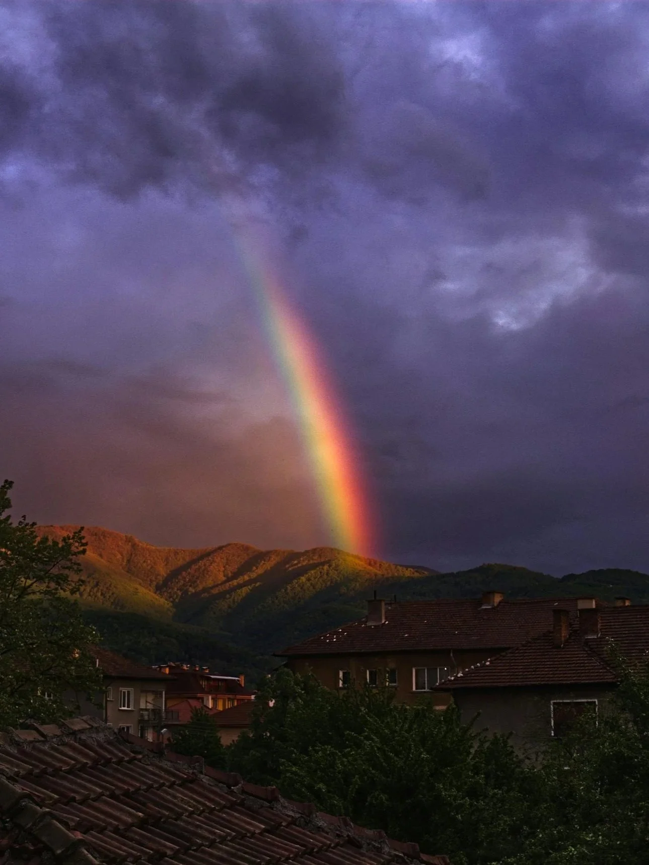 A vibrant rainbow arching over a hilly landscape with houses in the foreground and dark storm clouds in the sky.