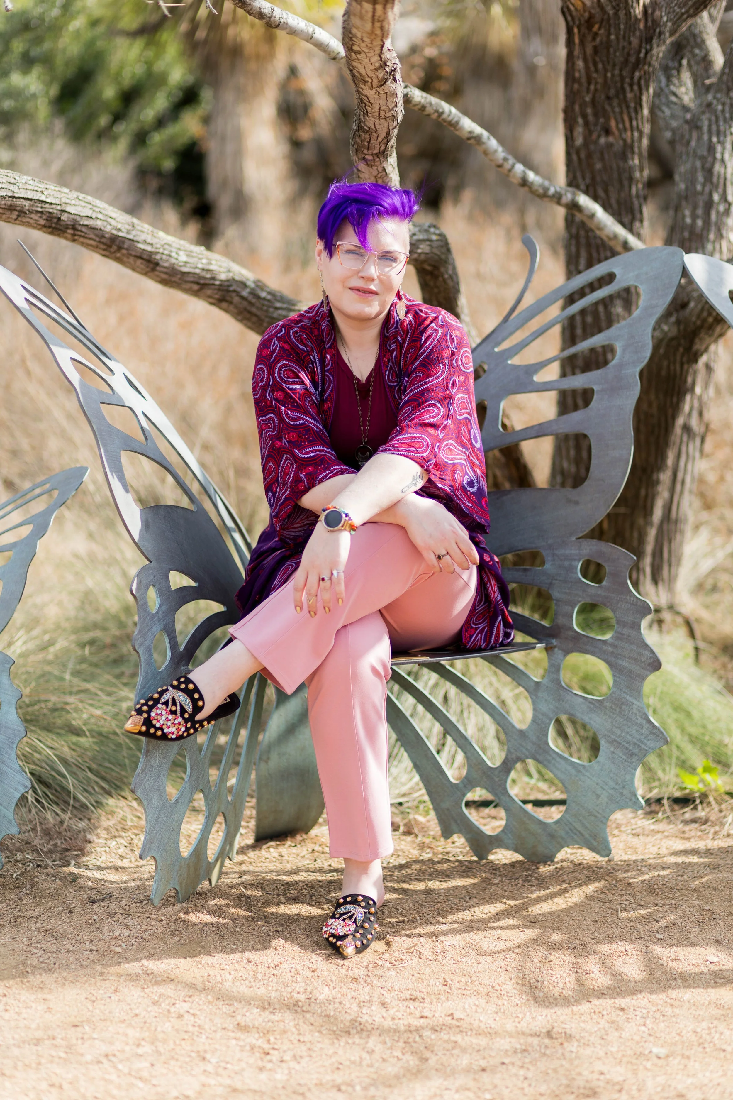 A woman with purple hair and glasses sitting on a butterfly-shaped metal bench in an outdoor setting with trees in the background.