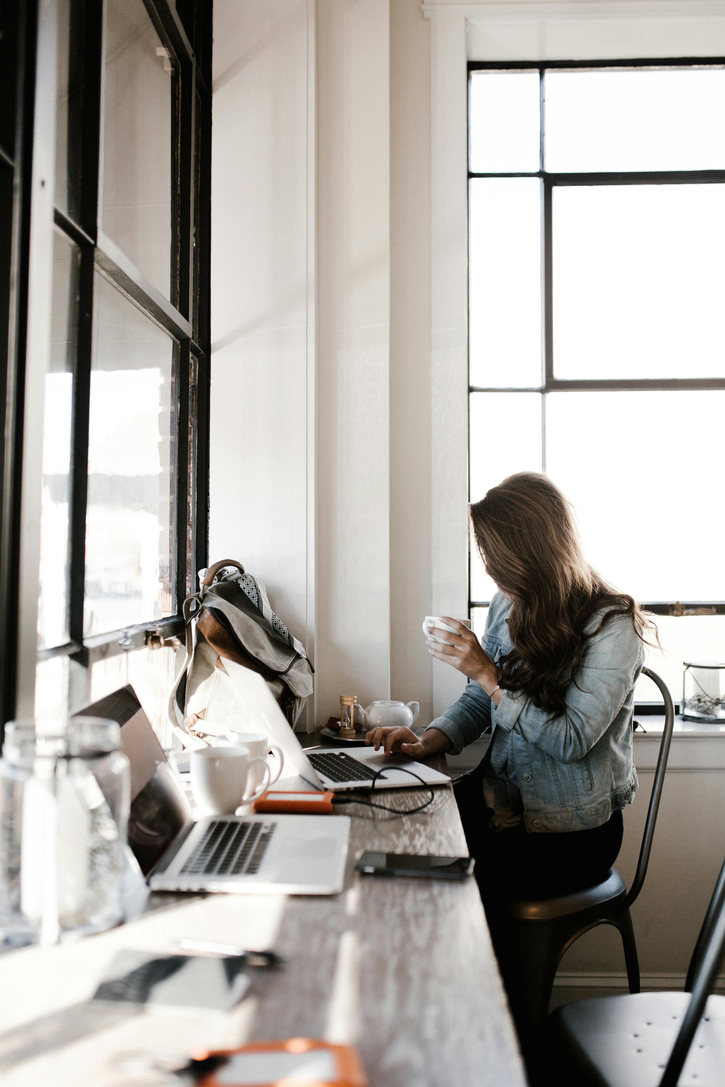 A woman sitting at a desk in front of a large window, looking at her phone with her laptop, coffee cup, and backpack on the desk.