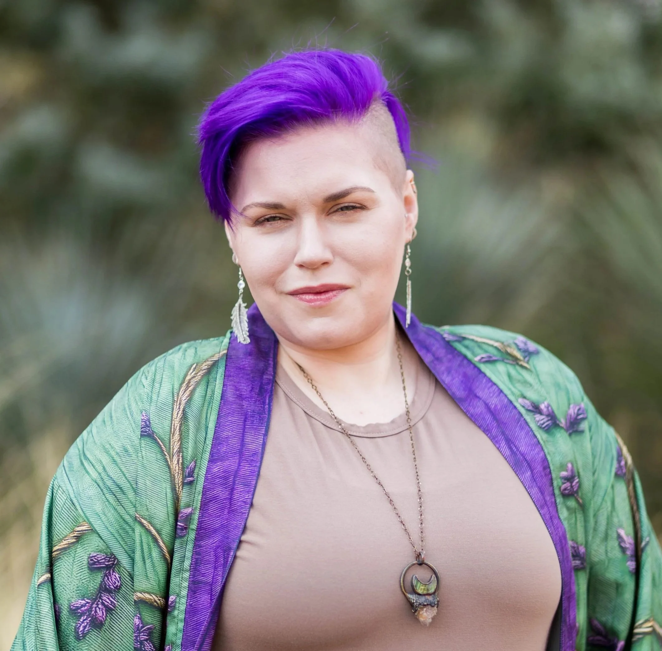 Close-up of a woman with purple hair styled in an undercut, wearing earrings, a necklace, and a colorful scarf, outdoors with a blurred green background.