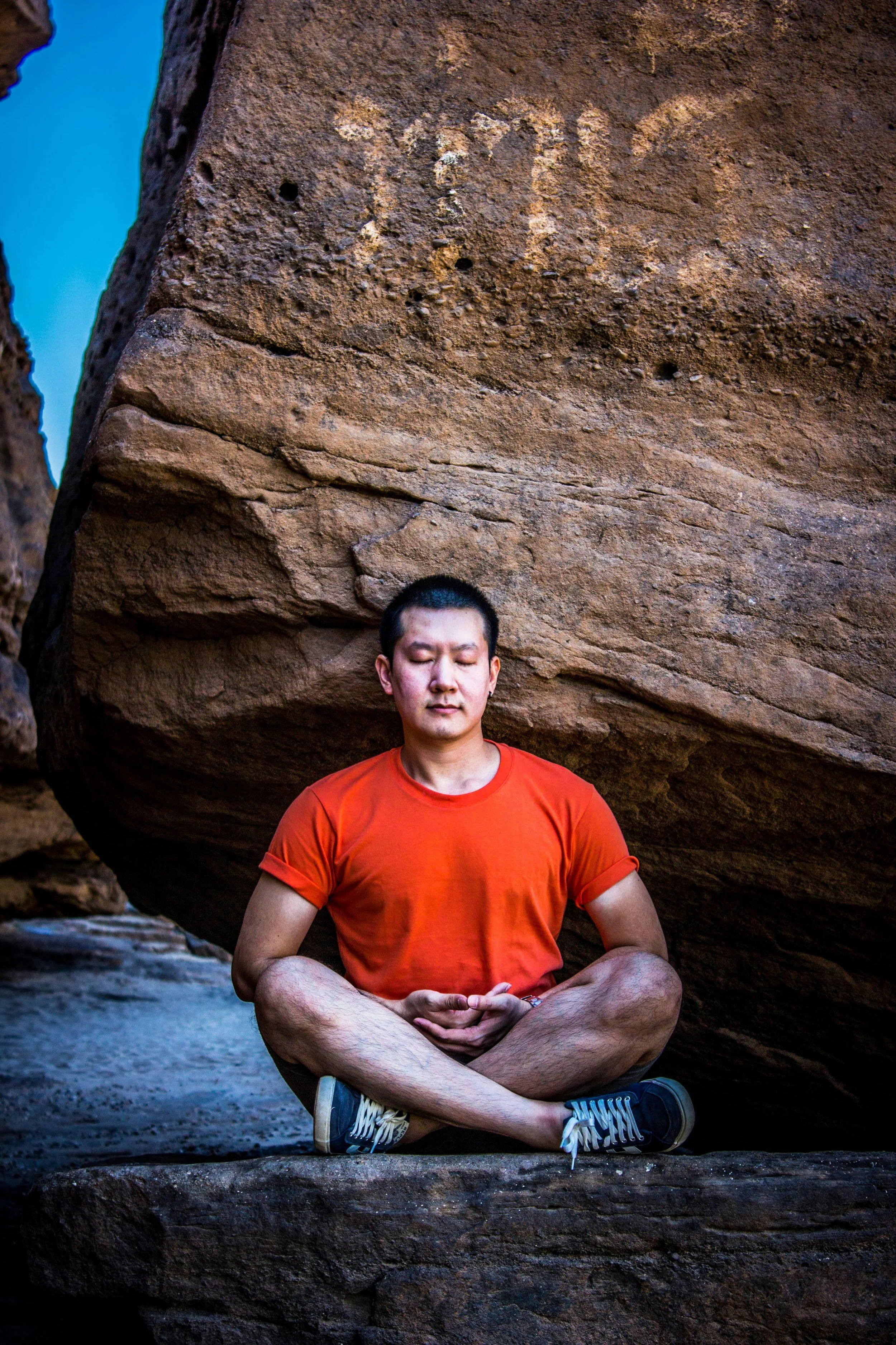 A man practicing yoga or meditation in a seated lotus position outdoors, with a large rock formation behind him and the word "LOVE" faintly etched in the rock above his head.