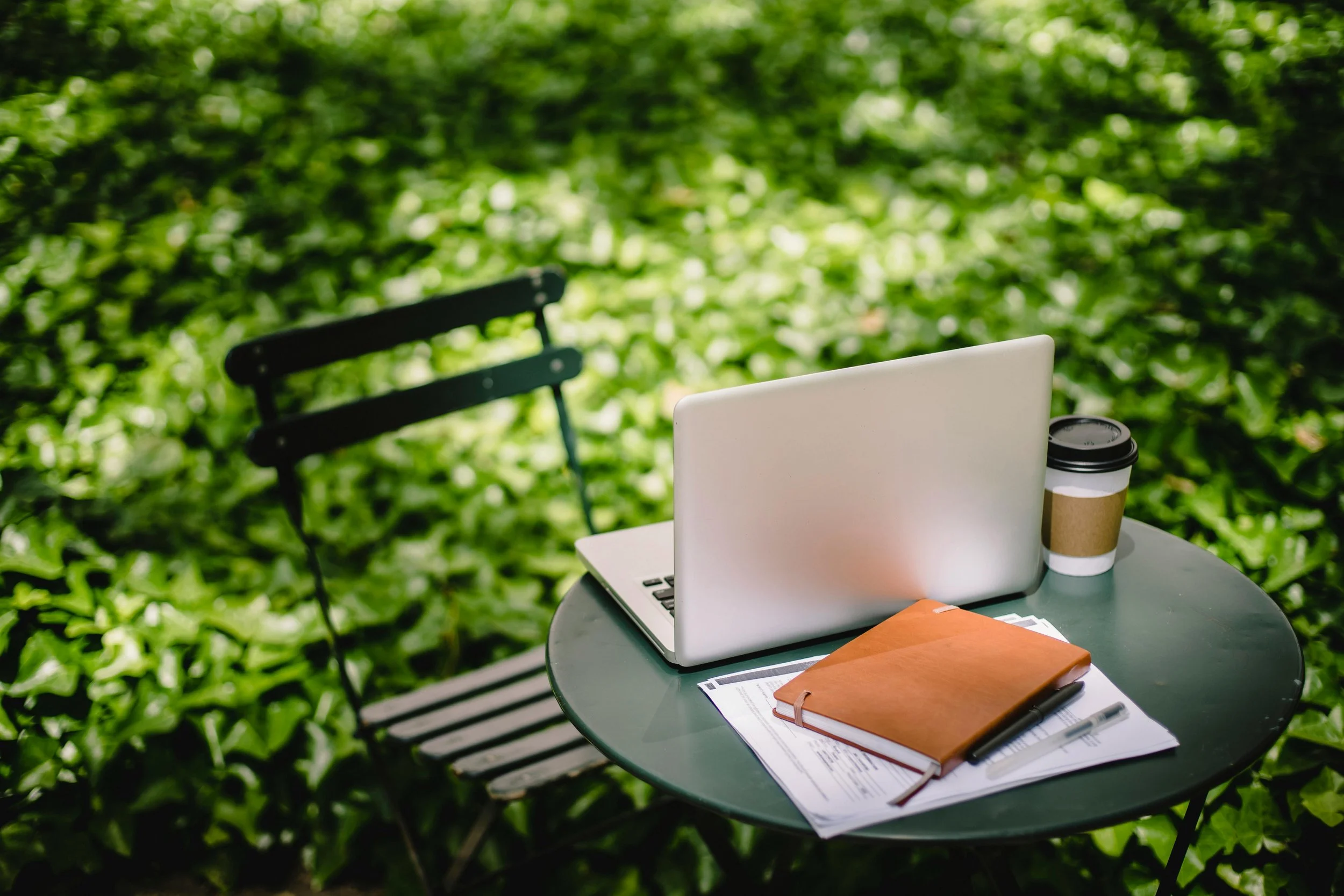 A round outdoor table with a laptop, a closed notebook, some papers, a pen, and a disposable coffee cup, set among lush green foliage.