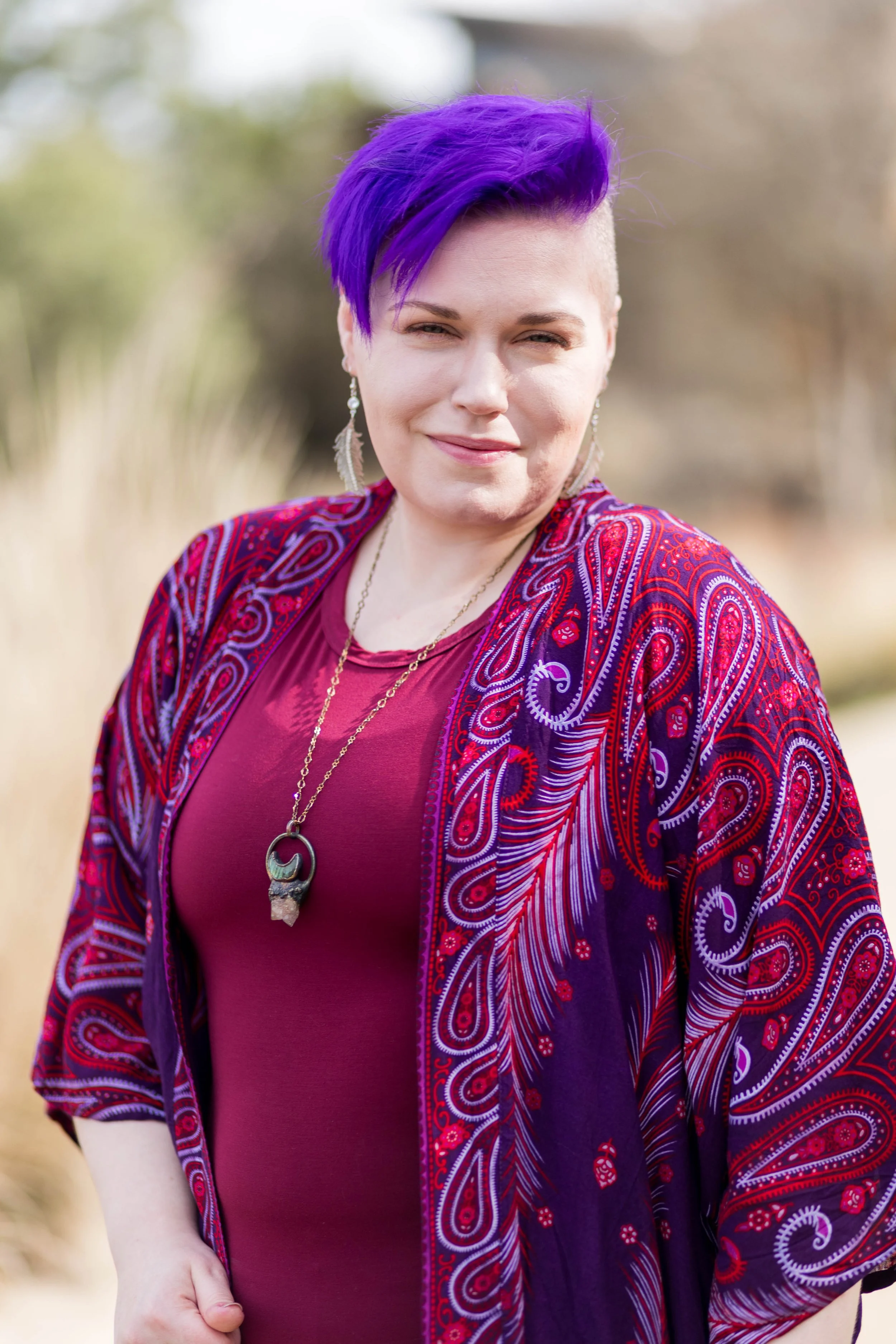A woman with purple hair, wearing a red top and a colorful, patterned purple and red shawl, standing outdoors.