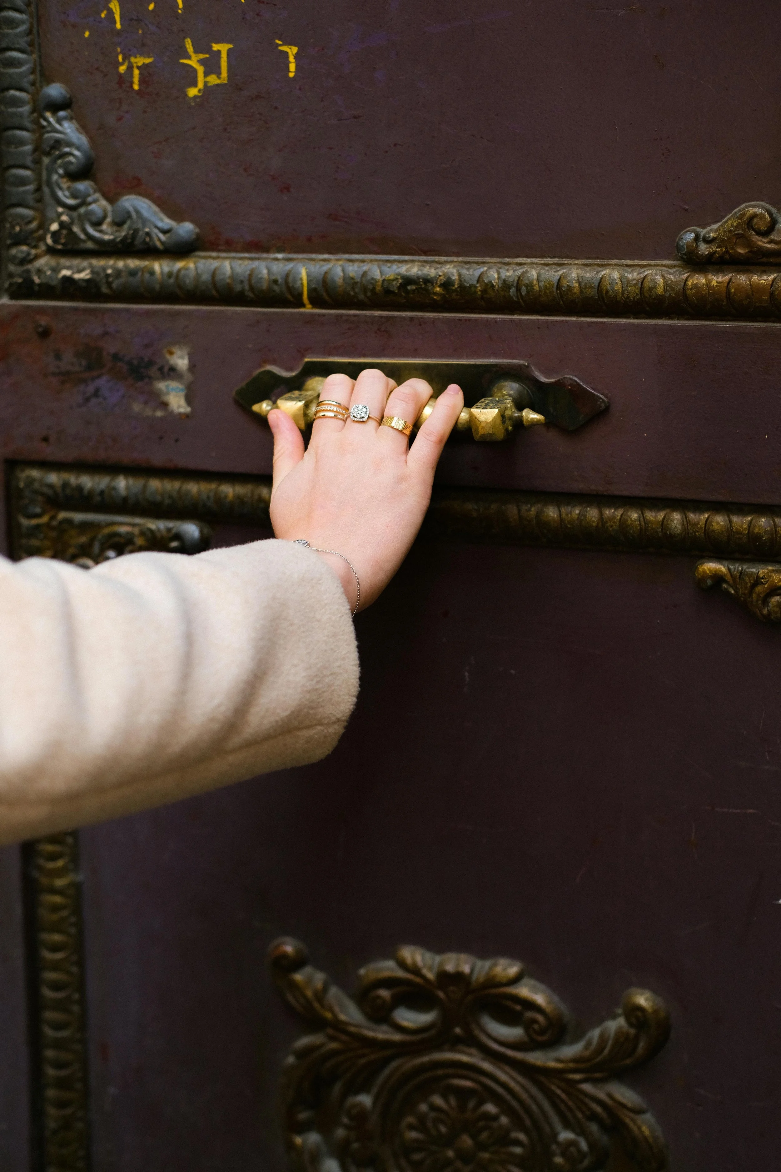 A person's hand with multiple rings touching a vintage brass handle on an ornate dark wooden piece of furniture.