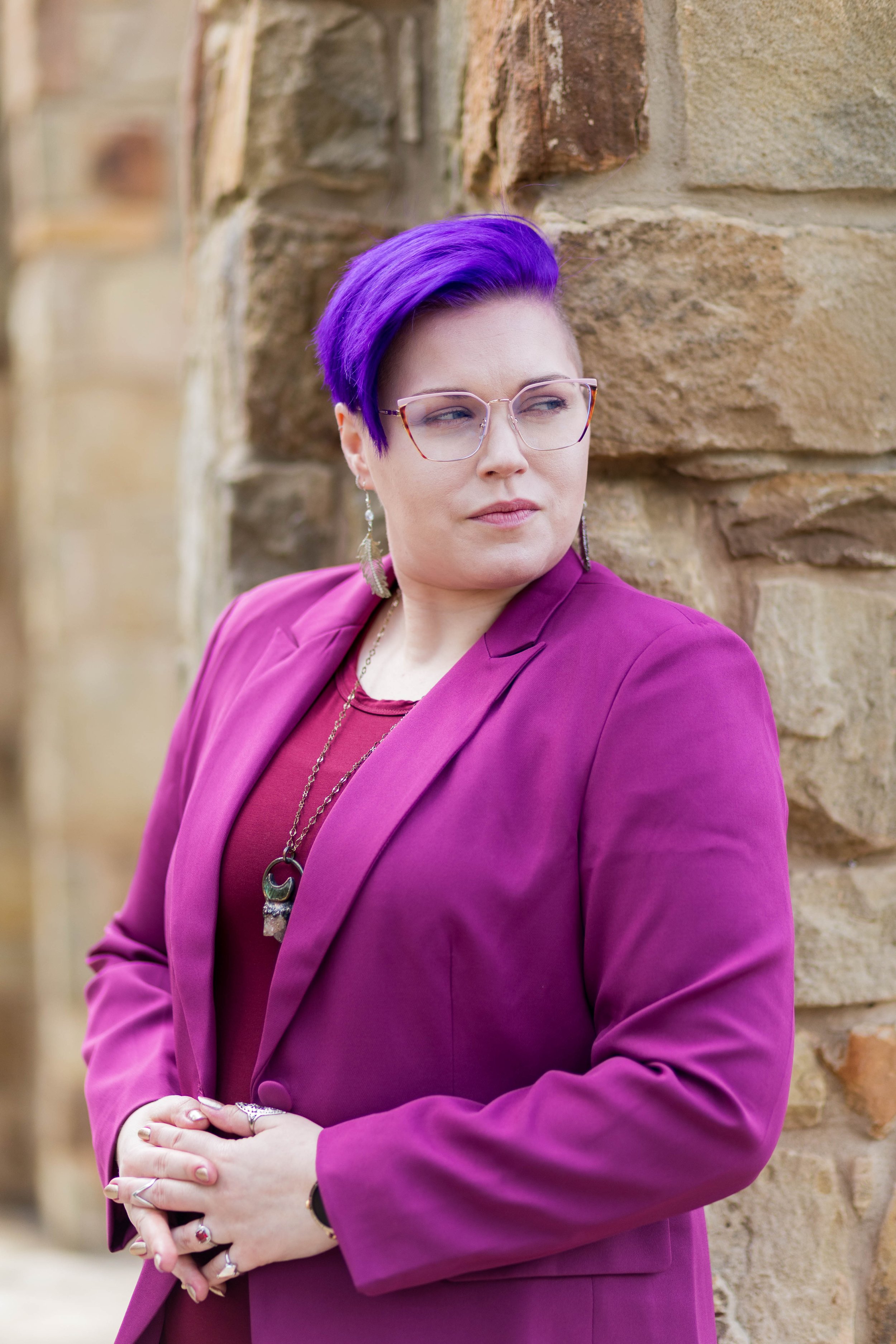 A woman with purple hair and glasses leaning against a stone wall, wearing a magenta blazer and jewelry.