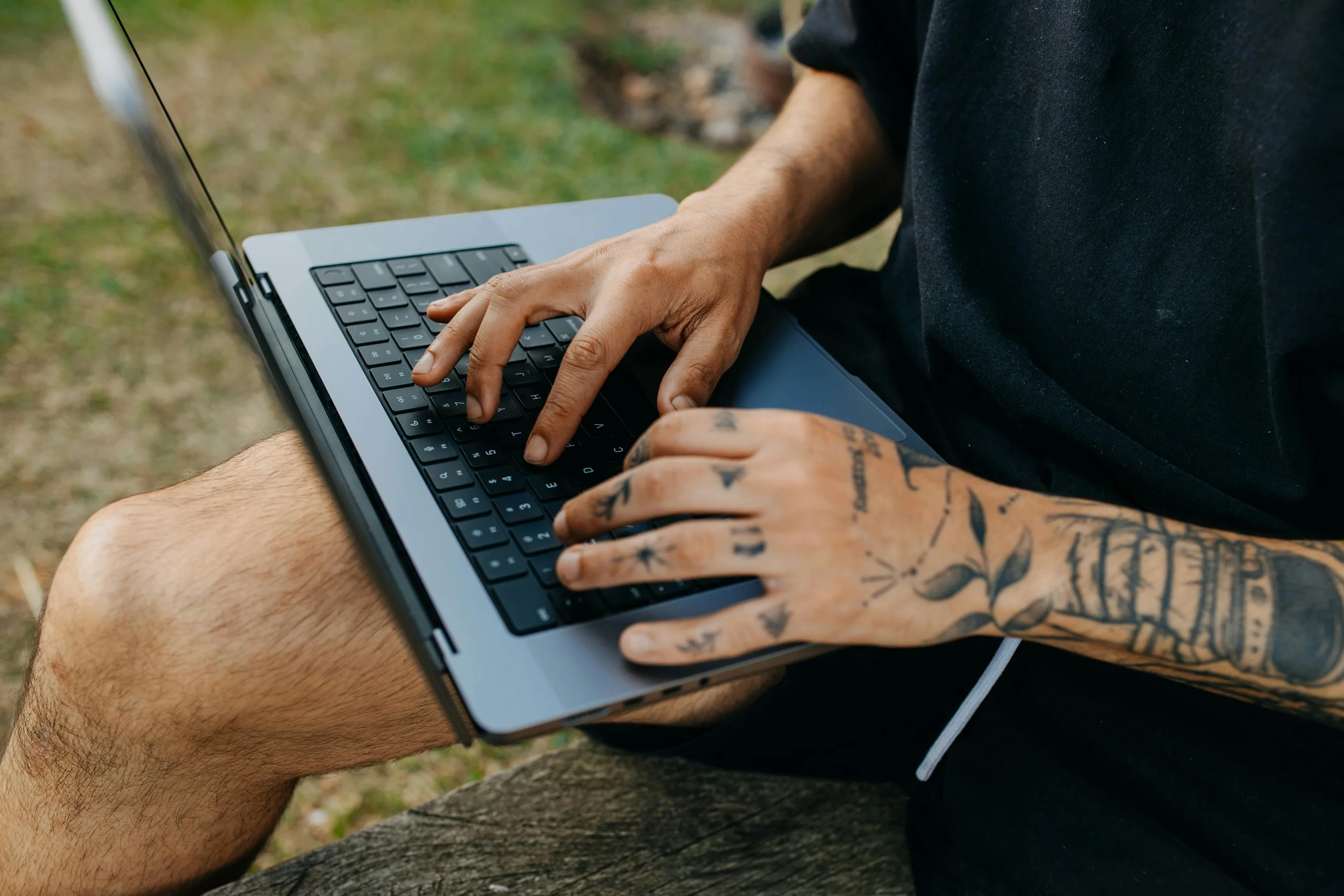 Person with tattoos using a laptop outdoors, sitting on a bench.
