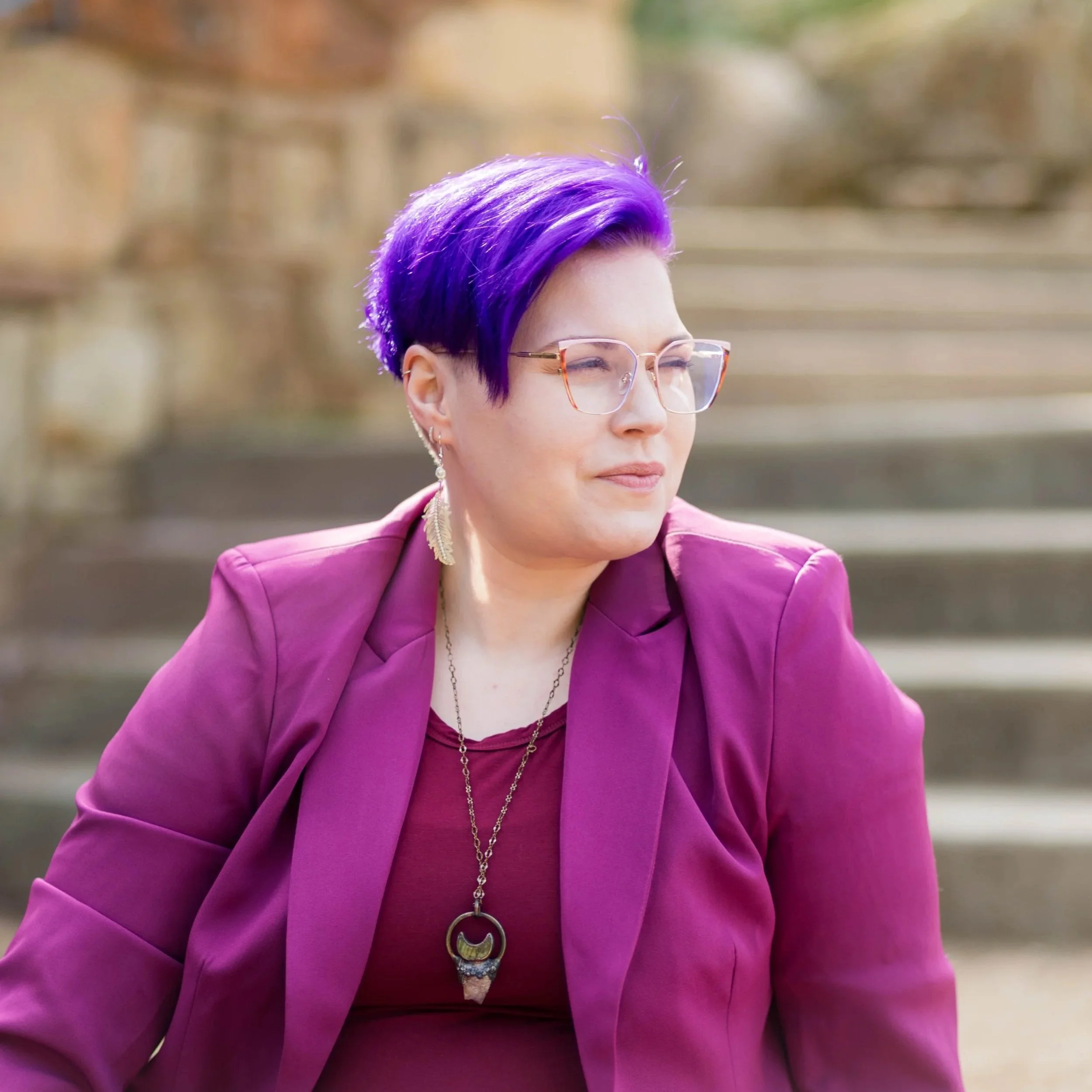 A woman with purple hair styled in a trendy cut, wearing glasses, earrings, and a purple blazer, sitting outdoors with a blurred background.