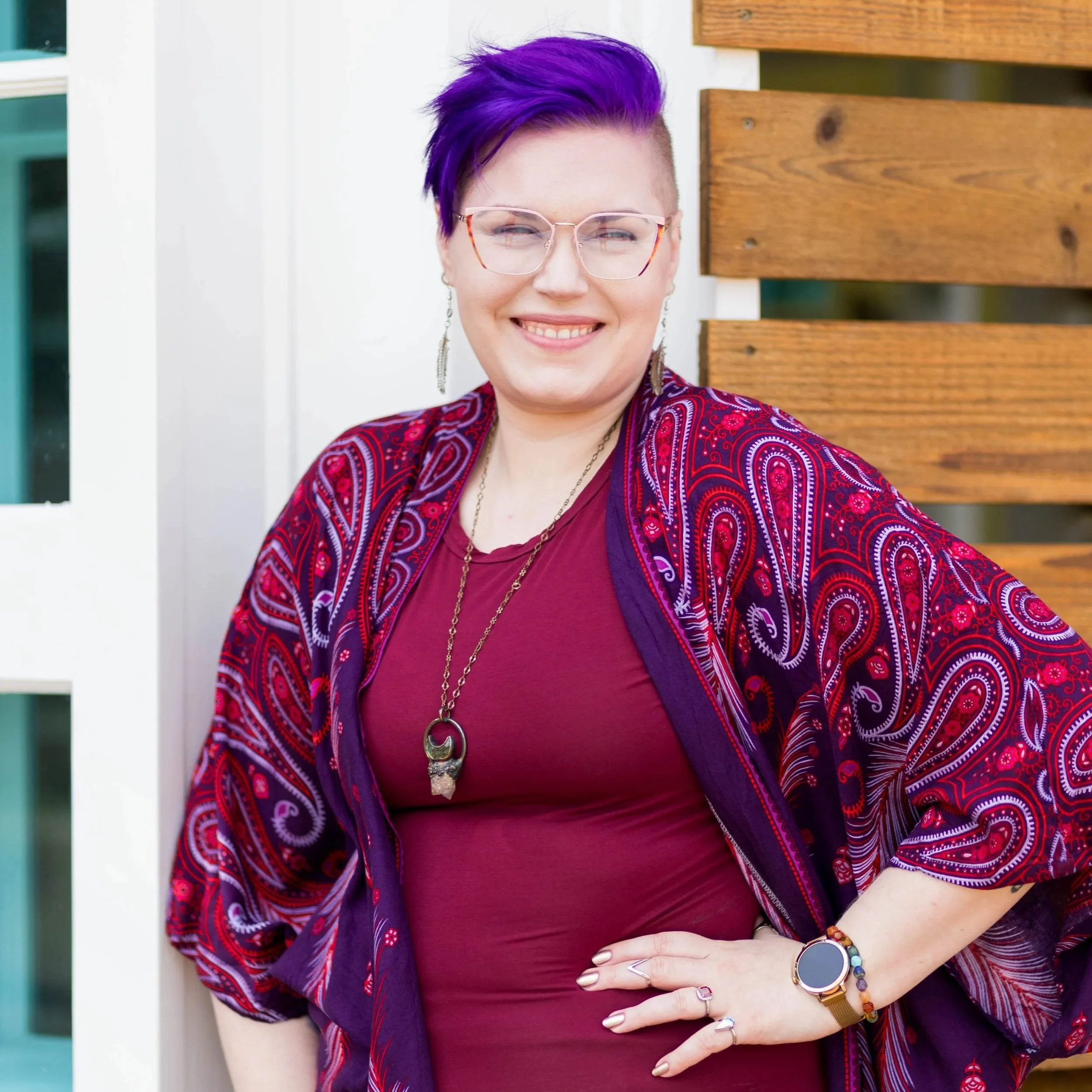 A woman with short purple hair, wearing glasses, earrings, a patterned red and purple jacket, and a purple shirt, smiling and holding her arm across her stomach outside near a white wall and wooden panel.