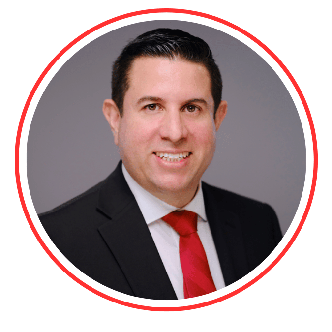 A professional headshot of a man with dark hair, wearing a black suit, white shirt, and red tie, smiling at the camera against a gray background.