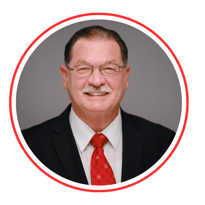 A middle-aged man wearing glasses, a black suit, white shirt, and red tie, smiling in front of a plain gray background.