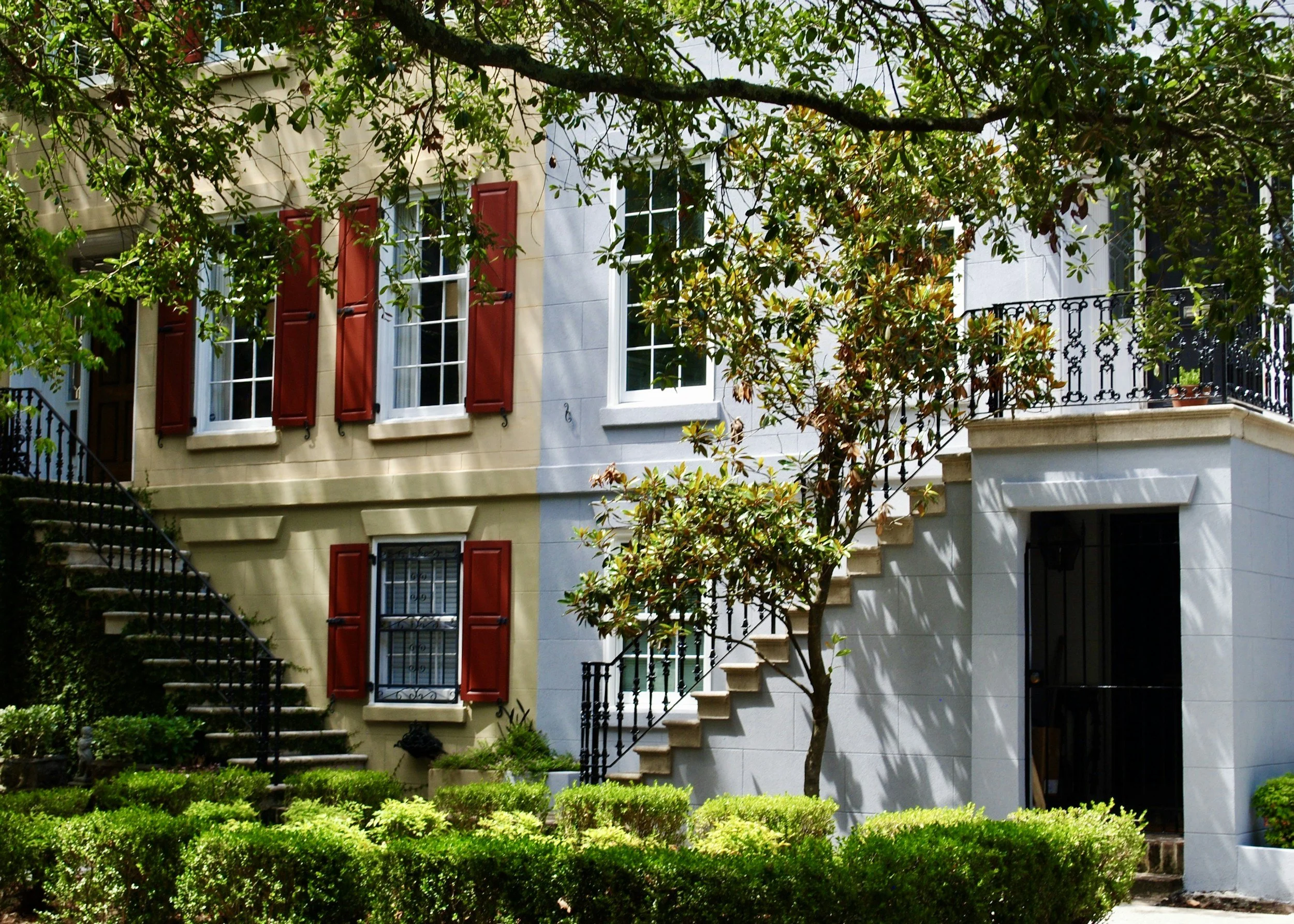 A two-story residential building with beige and gray walls, red window shutters, black wrought iron stairs, and lush greenery including trees and bushes near savannah college of art and design