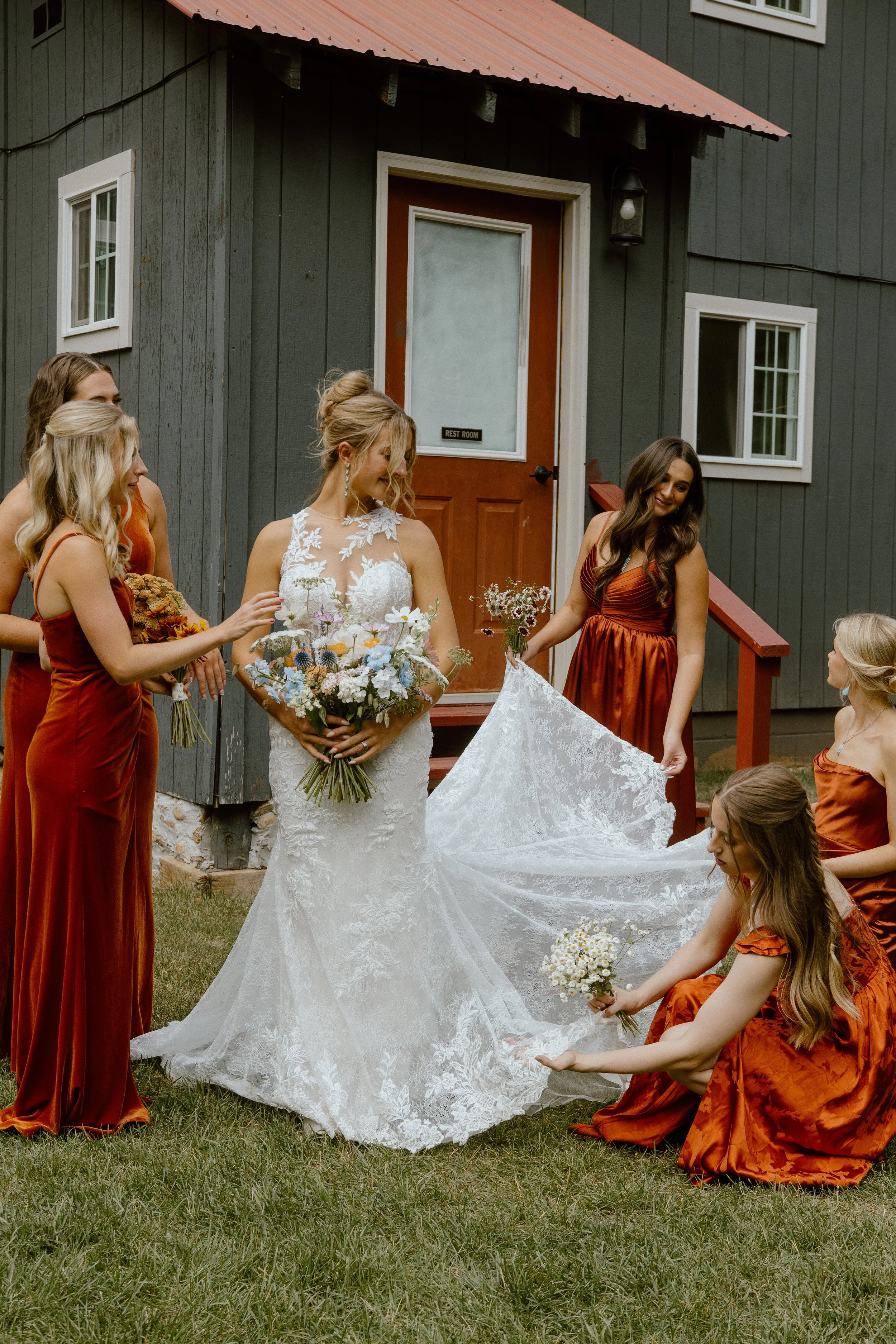 Bride in white lace wedding dress holding a bouquet of flowers with six bridesmaids surrounding her, dressed in rust-colored dresses, outside a dark green building with a small porch and window, as they prepare the dress, possibly for a wedding.