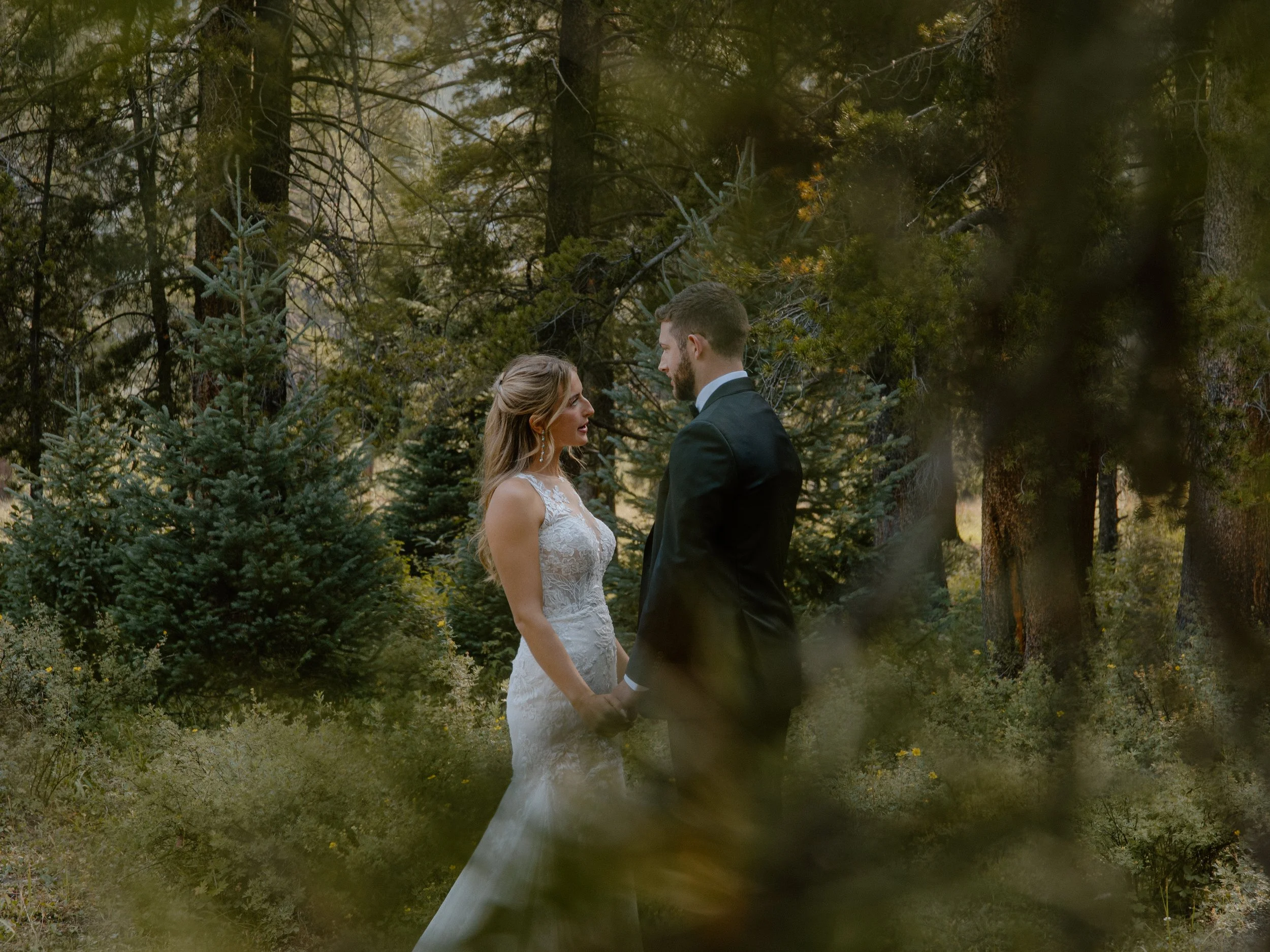 A bride and groom holding hands and facing each other in a forest, with trees and foliage around them.