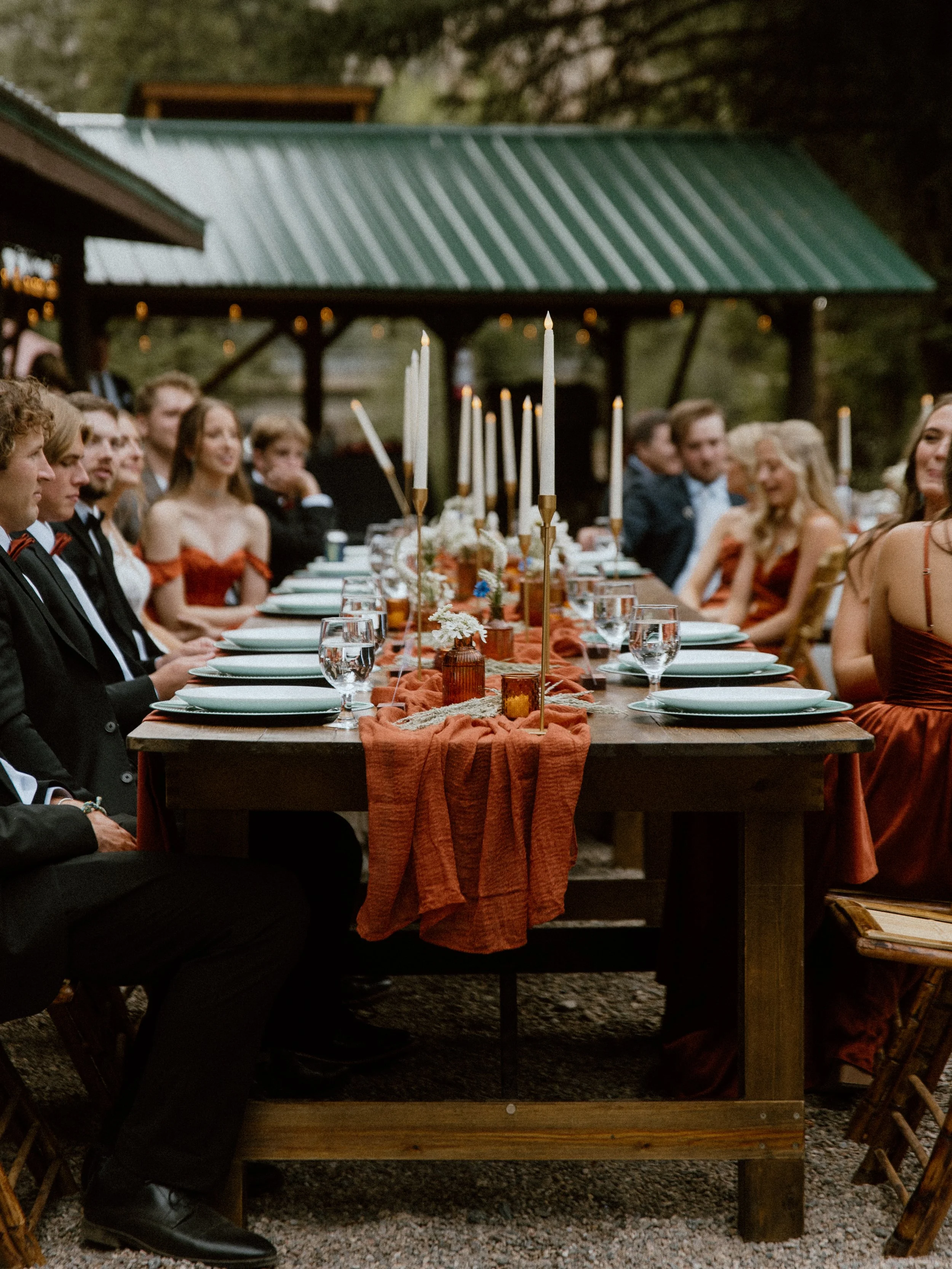 People dressed formally sitting at a long outdoor banquet table with candles, flowers, and a rustic setting.