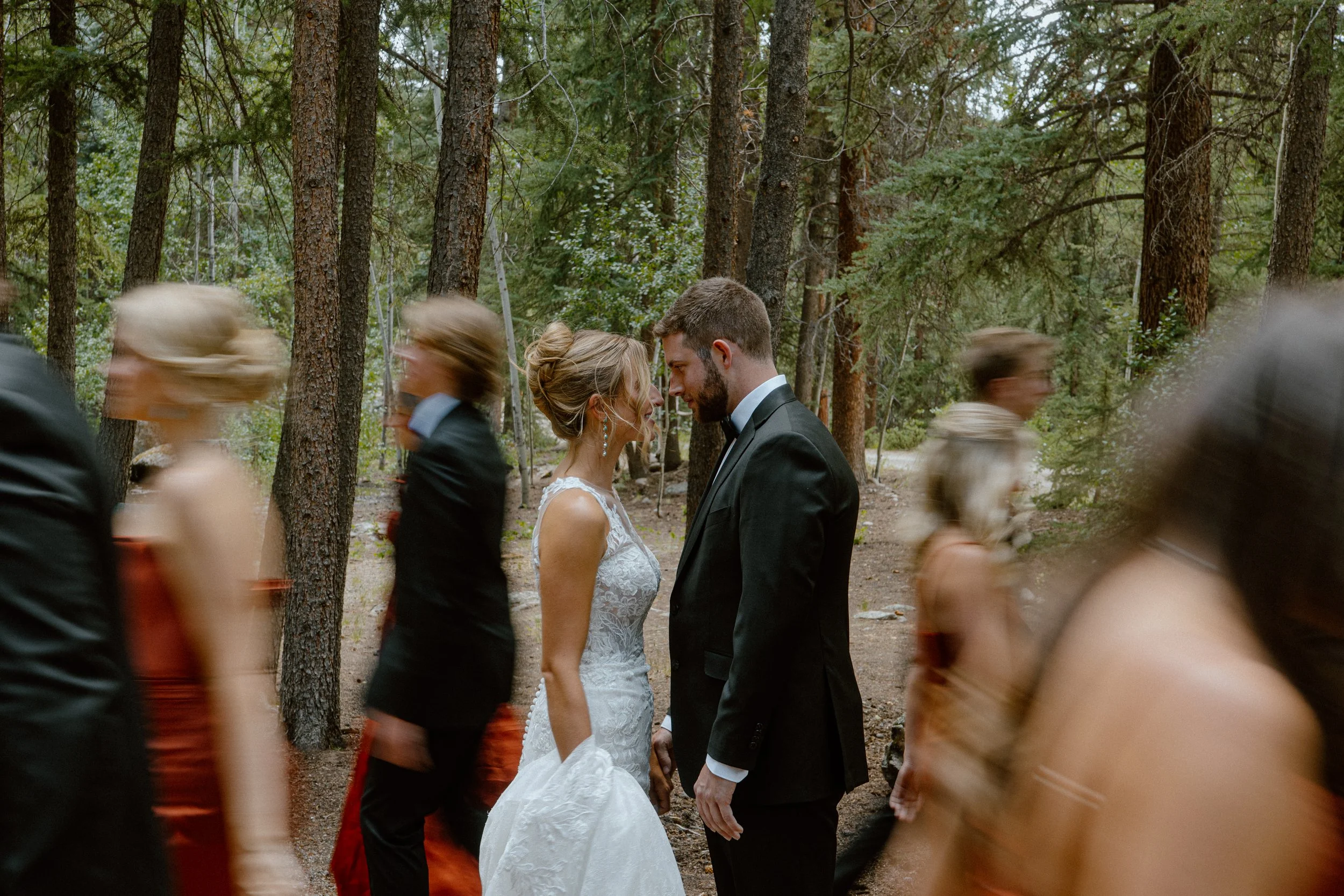 A bride and groom face each other in a forest, surrounded by blurred wedding guests walking by.