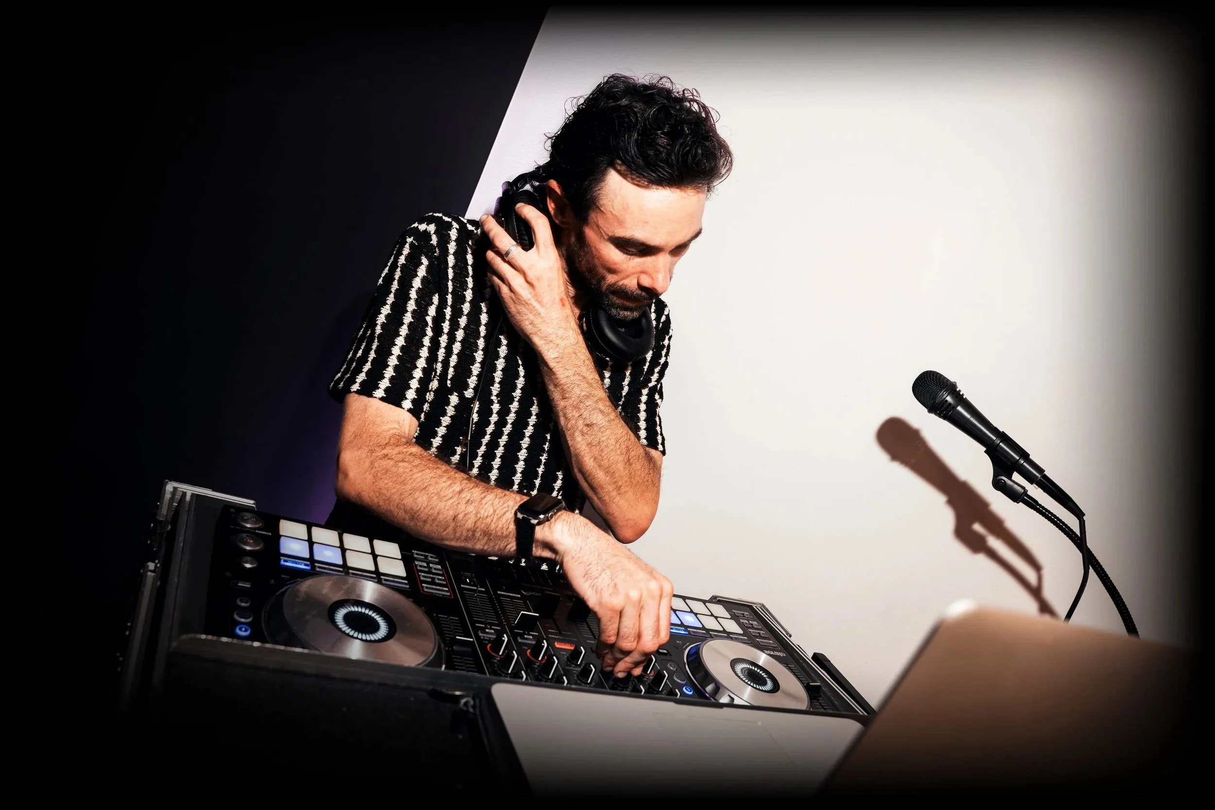 A man DJing with headphones, operating a DJ controller and turntables in front of a microphone on a black stand, with a plain white background.