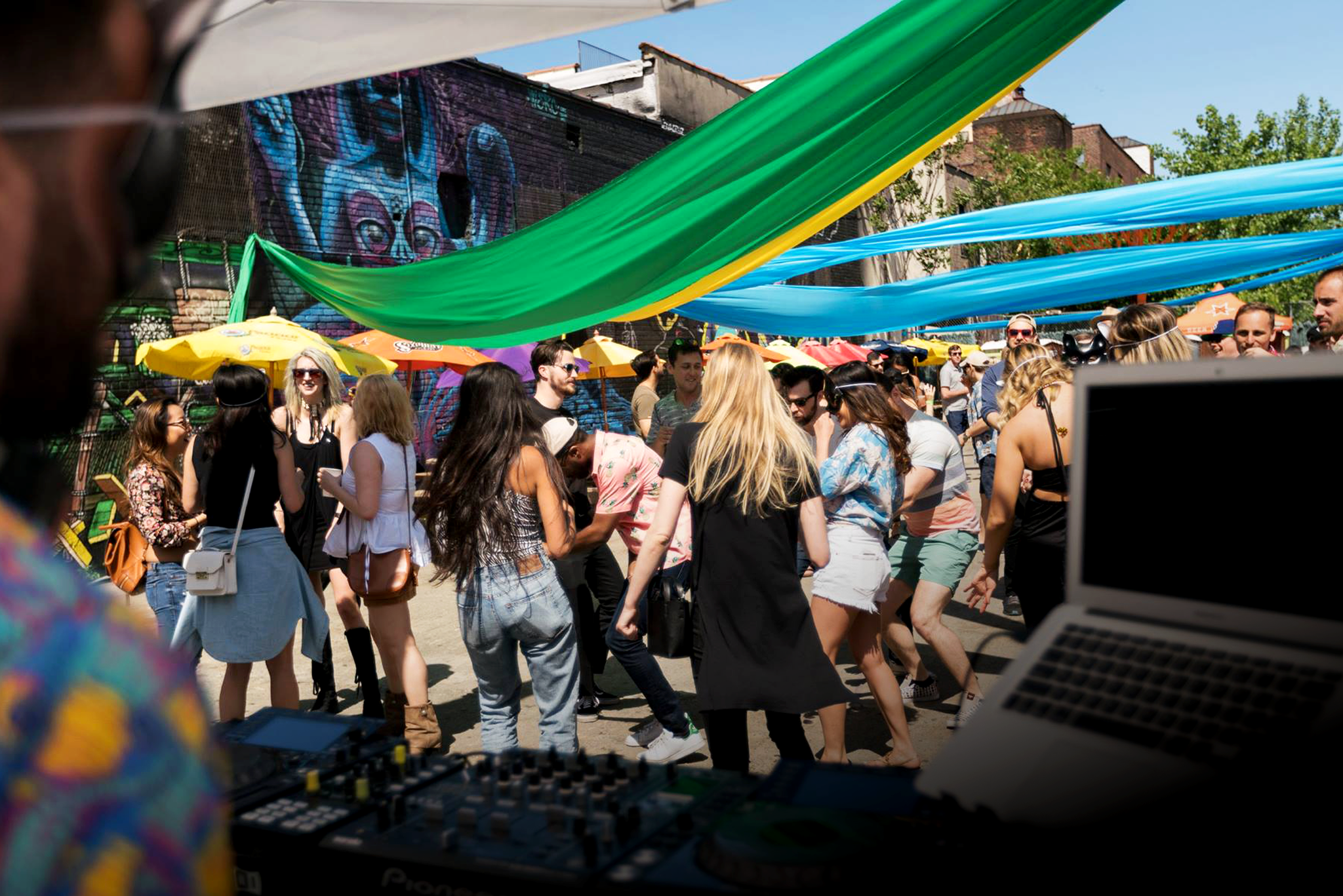 Crowd of people at an outdoor event with colorful streamers overhead, a graffiti wall, and umbrellas for shade. A DJ or musician is playing music in the foreground.
