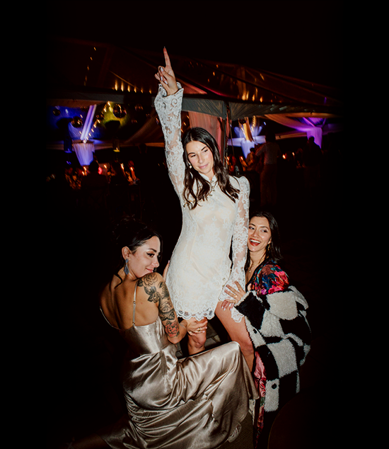 Three women celebrating at a party with colorful lights; one woman is standing on a chair with her arm raised, dressed in a white lace dress, while the other two women are seated and smiling.