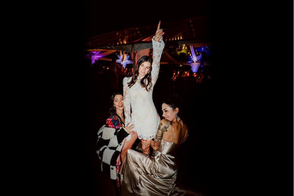 Three women dancing at a party, one standing on a chair with her arm raised, one smiling in colorful clothing, and the third with tattoos in a satin dress, all enjoying the celebration in a brightly lit venue.
