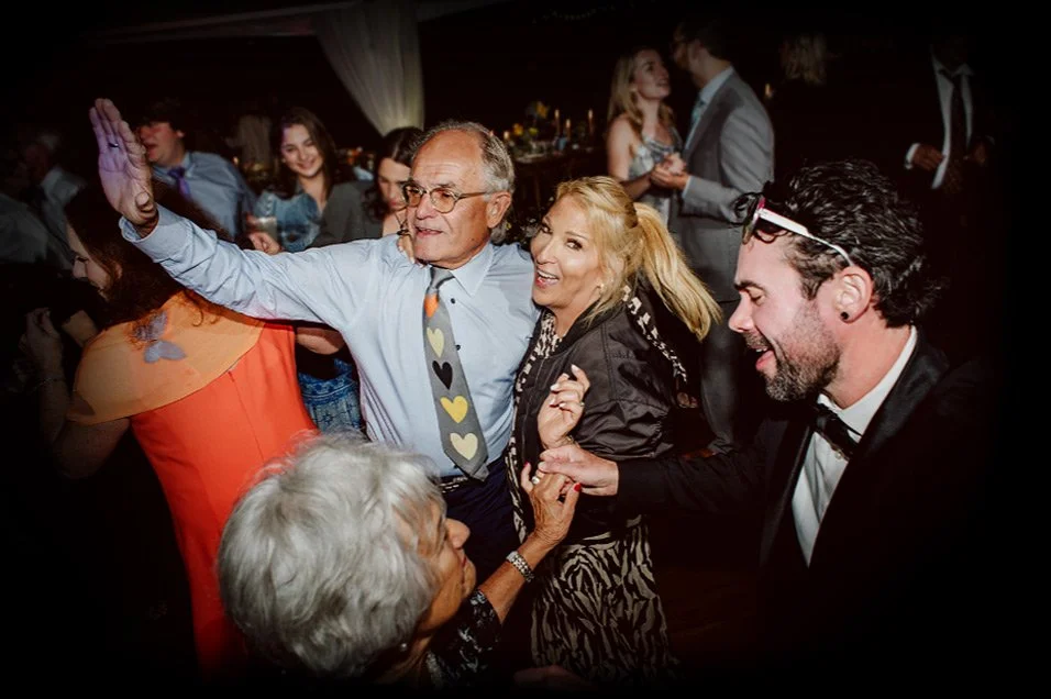 People dancing and enjoying themselves at a lively party or celebration, with a man in glasses and a colorful heart tie, a woman with blonde hair, and a man in a suit, among other guests.