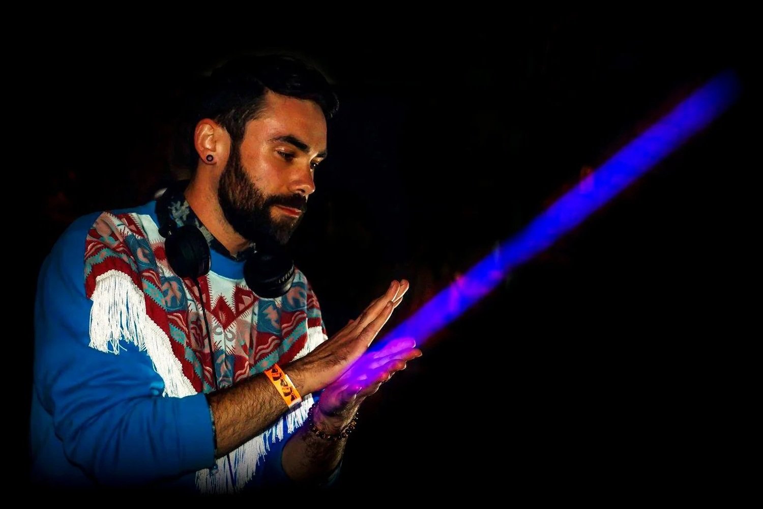 A DJ with a beard and styled hair, wearing a colorful fringed sweater, black headphones around his neck, and an orange wristband, is looking down with his hands raised, in front of a dark background with a purple laser beam.