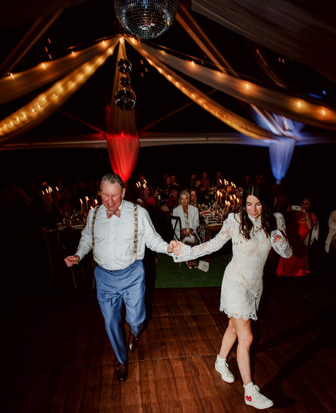 A man and woman dancing together at a celebration or wedding reception under a decorated ceiling with hanging disco balls and string lights, with guests seated at tables in the background.