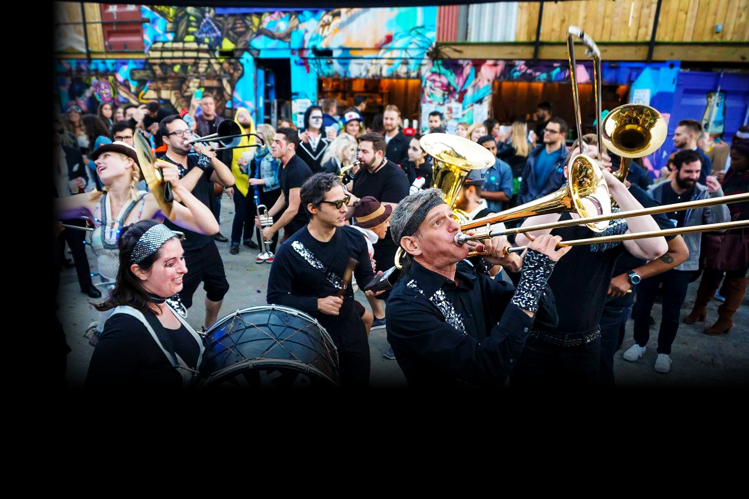 A lively street performance with musicians playing brass instruments, drums, and singing, surrounded by a crowd of onlookers, graffiti-covered wall in the background.