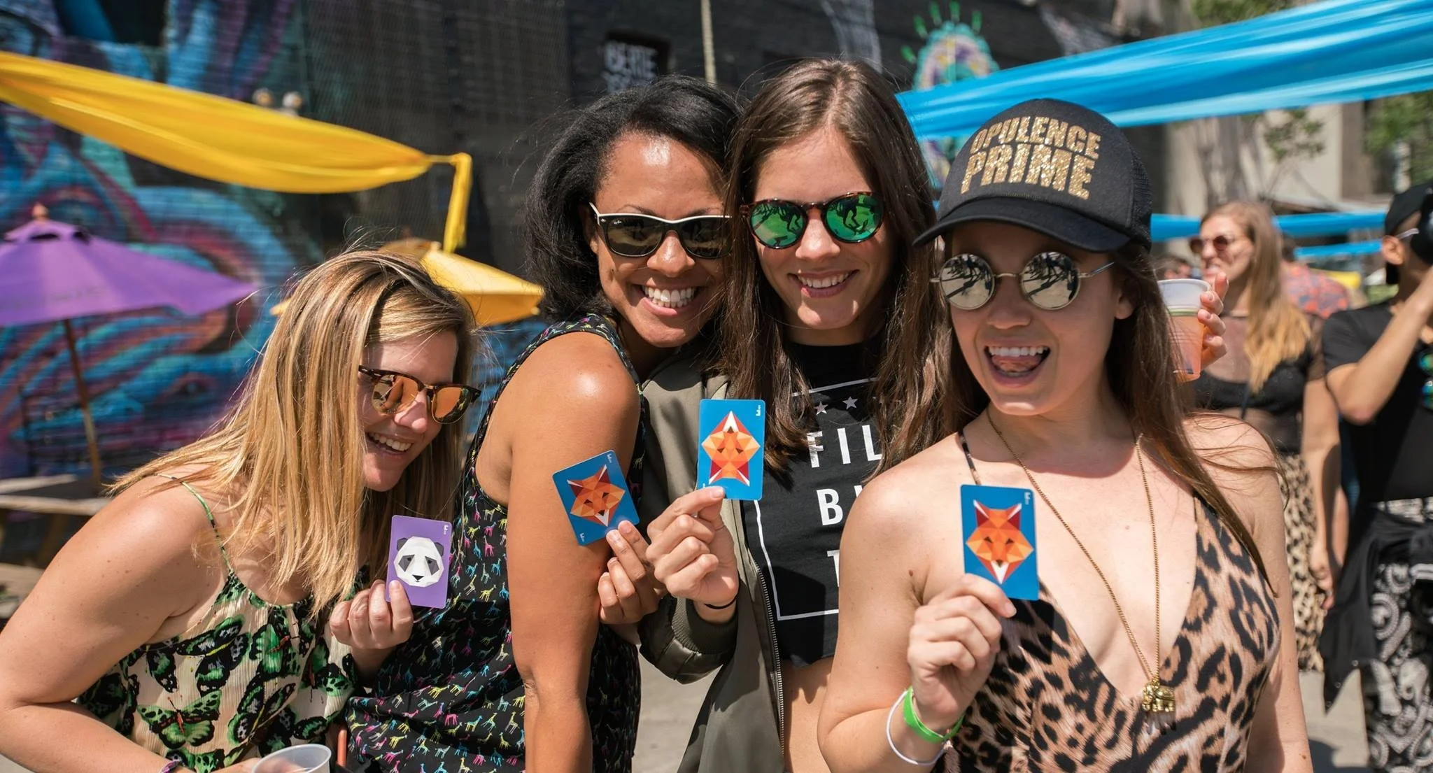 A group of four women smiling, holding colorful cards with animal logos, at an outdoor event with decorated background and other people in the distance.