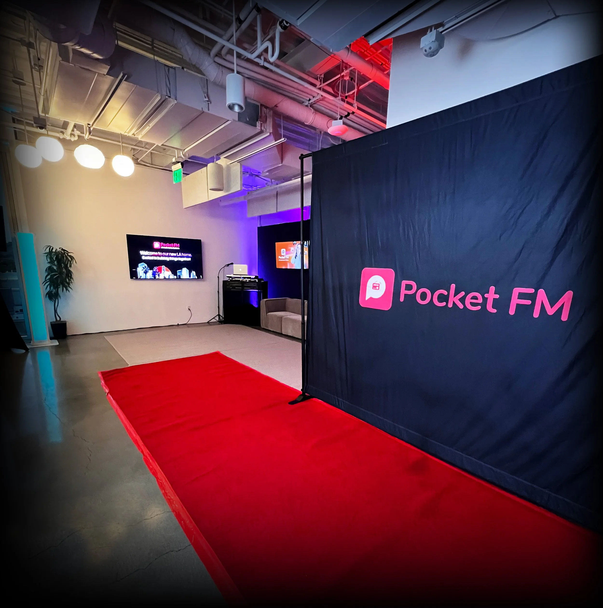 Indoor setting with a red carpet leading to a stage area, with black curtains and television screens displaying a welcome message for Pocket FM, purple and blue lighting, ceiling with exposed pipes and ventilation, and a few seating chairs.