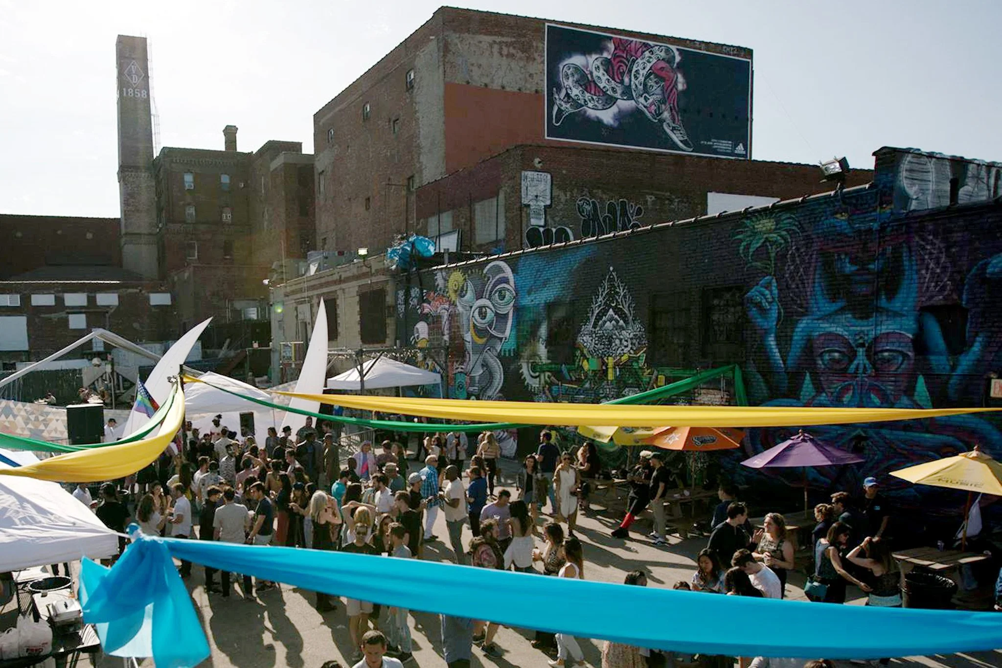 A crowded outdoor event with people walking and gathering under yellow and green canopy shades. Graffiti art decorates brick buildings in the background, along with murals and street art, including a snake and a face with glasses. Tents and booths are set up, suggesting a festival or street fair.