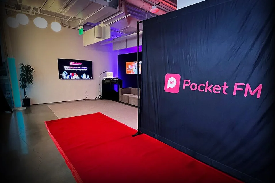 Indoor event space with a red carpet, a Black backdrop with pink 'Pocket FM' logo, a seating area with a beige sofa, television screens, and some plants.