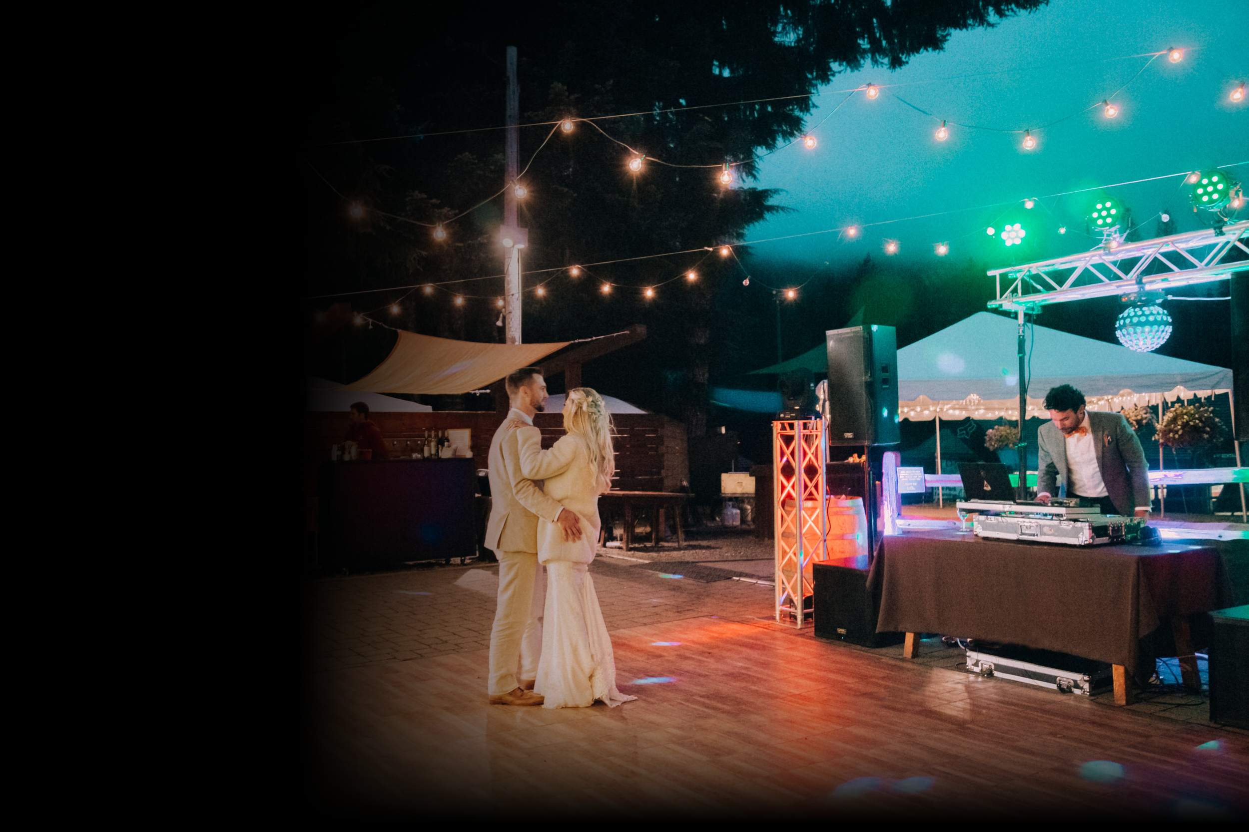 A newlywed couple dancing at their wedding reception outdoors during evening, with string lights and DJ equipment nearby.
