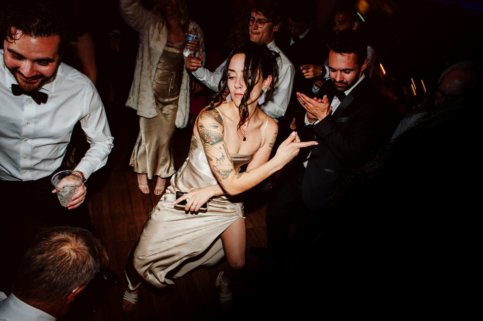 People dancing at a party, with a woman in a silver dress with tattoos on her arm in the center.