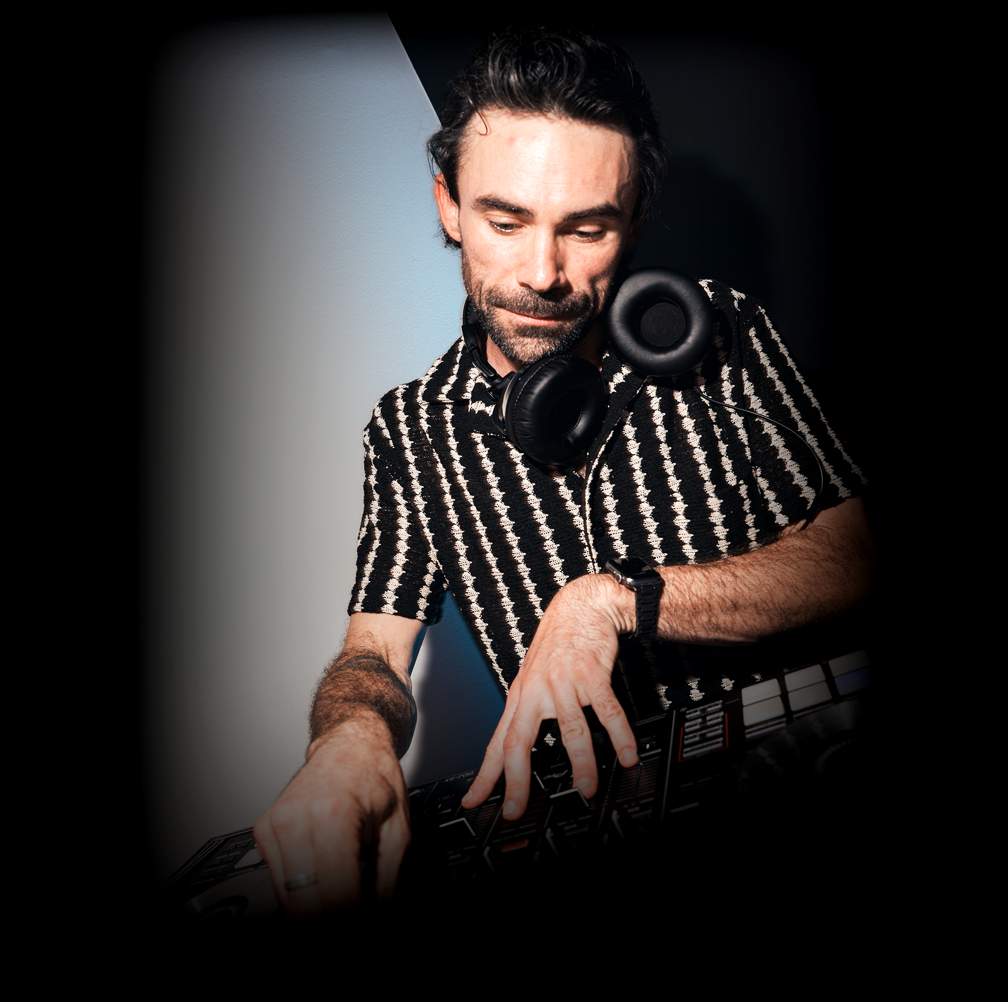 A man with dark hair and a beard wearing a patterned shirt, headphones around his neck, and a black watch, working on a digital keyboard or synthesizer.