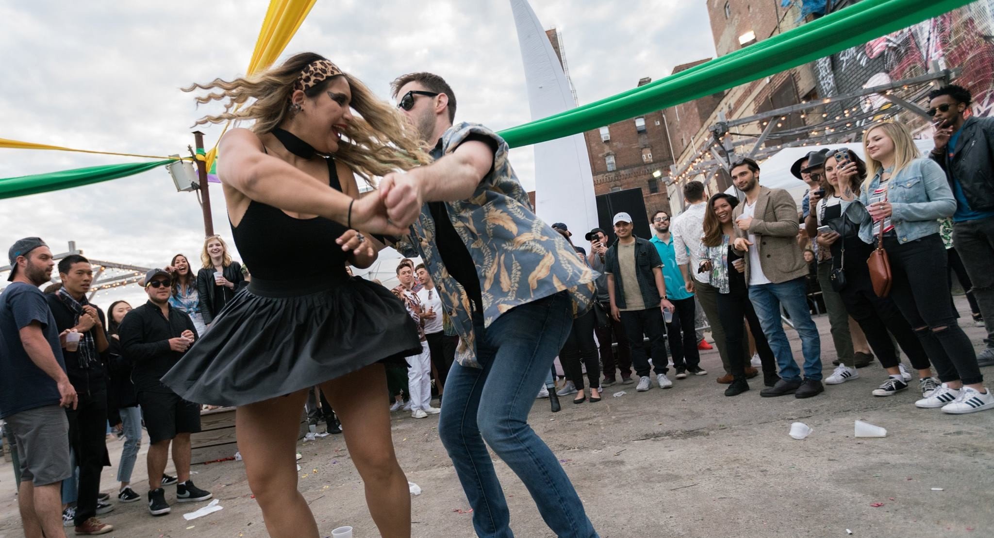 Two people dancing closely at an outdoor event while a crowd watches, with city buildings and string lights in the background.