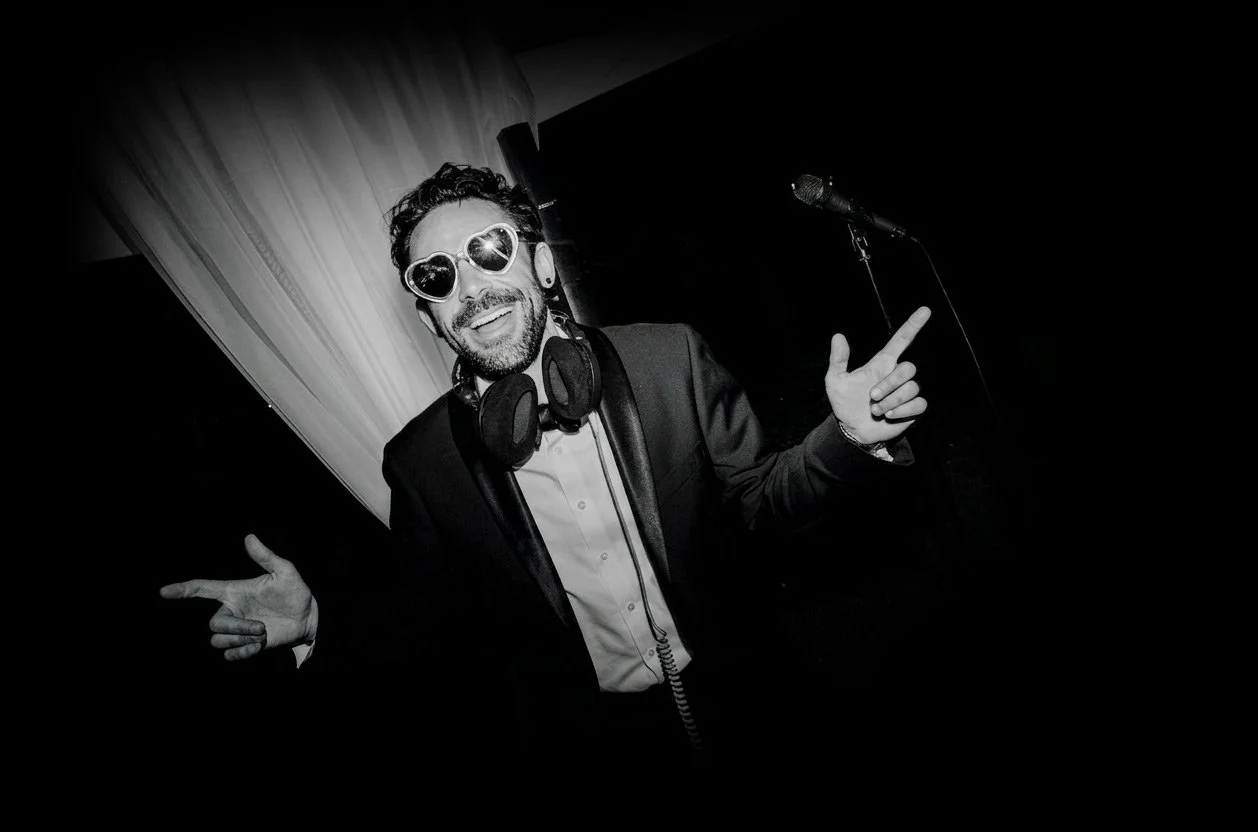 A man wearing heart-shaped sunglasses, a tuxedo with a white shirt, and headphones around his neck, smiling and pointing in different directions near a microphone on a stage.