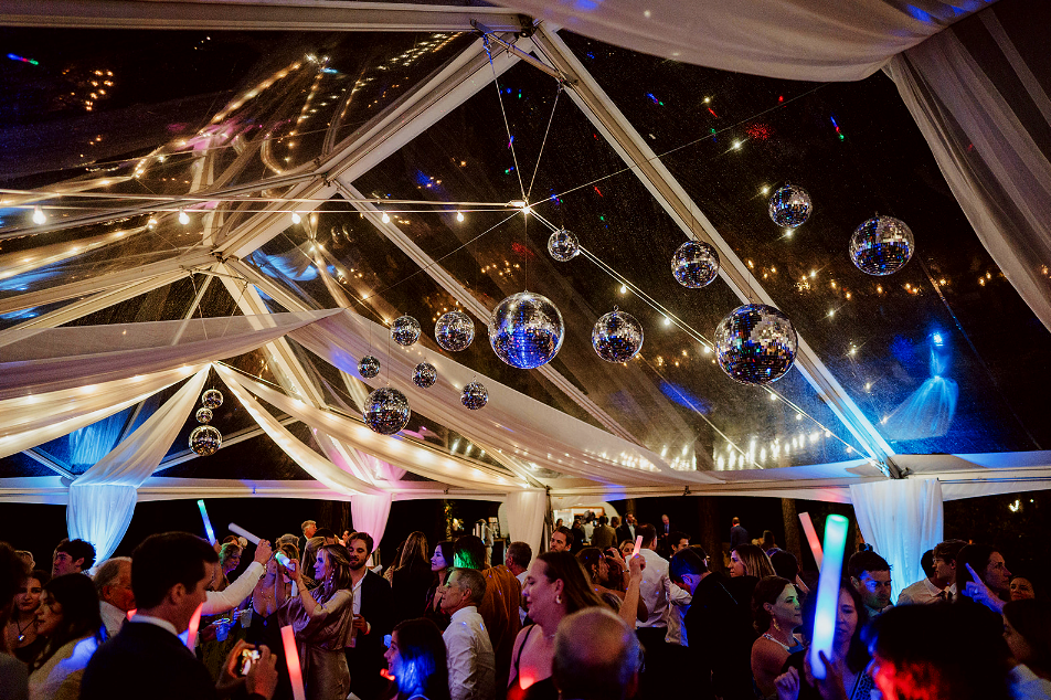 People dancing at a party under a decorated tent with hanging disco balls and string lights.