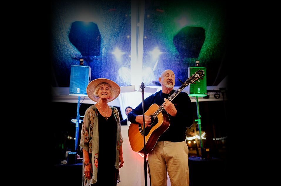 A man and a woman perform music on stage, with the man playing an acoustic guitar and singing into a microphone, while the woman stands beside him. The stage is set with speakers and bright colorful lights overhead.