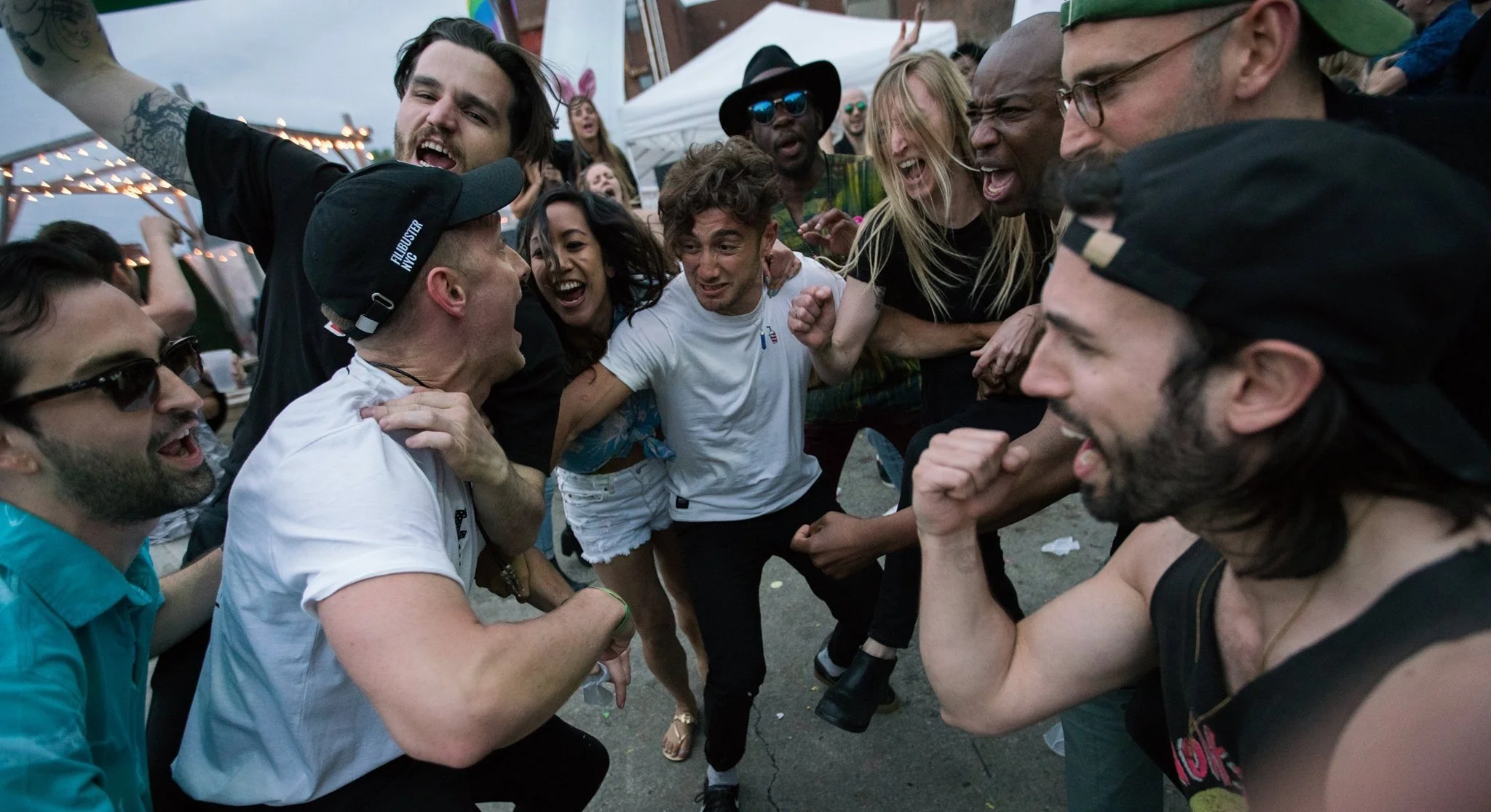 A group of diverse young adults at an outdoor party, engaged in a lively fistfight with expressions of excitement and intensity, under string lights and tents.