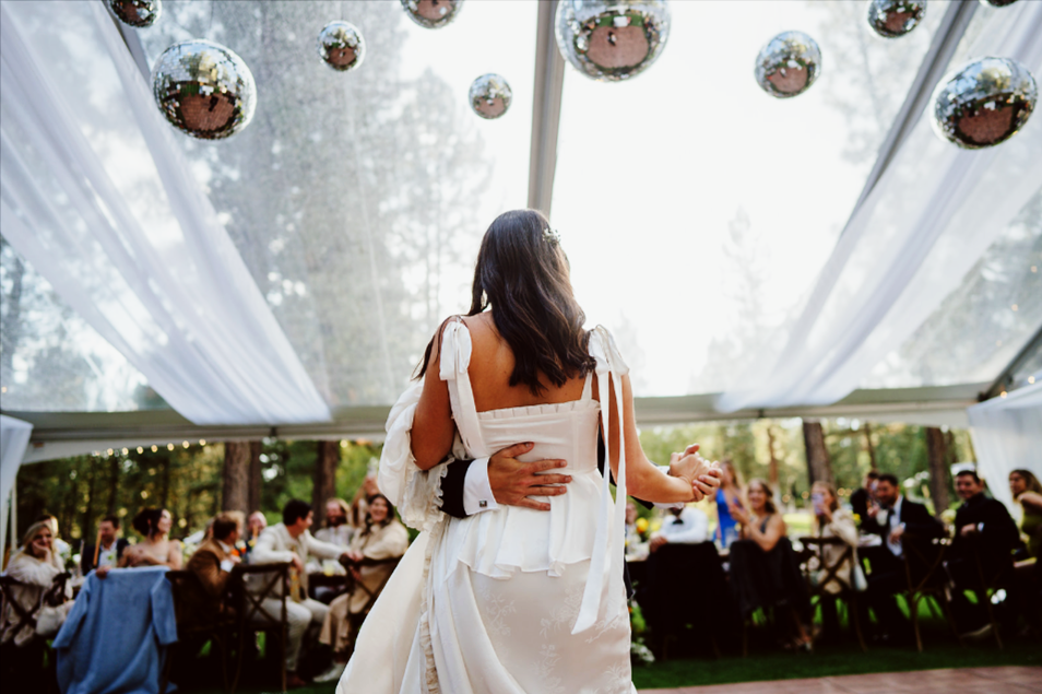 A bride and groom sharing a dance at their wedding reception, surrounded by seated guests under a tent with hanging metallic ornaments.