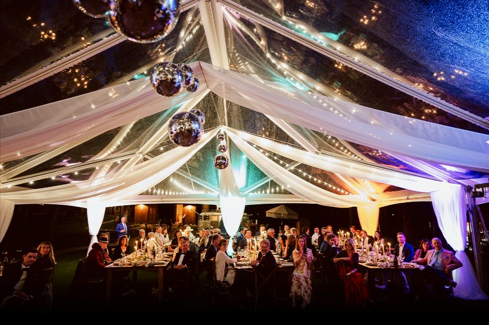 A large outdoor evening wedding reception under a decorated tent with hanging disco balls, string lights, and white drapery, with guests seated at long tables.
