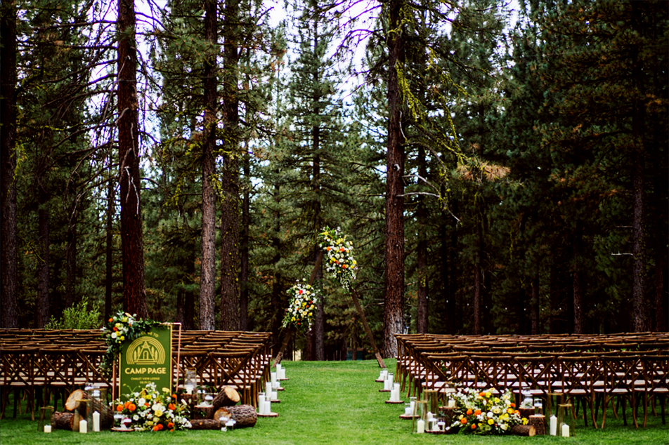 Outdoor wedding ceremony setup in a forest with rows of wooden chairs, floral arrangements, candles, and an arch decorated with flowers.