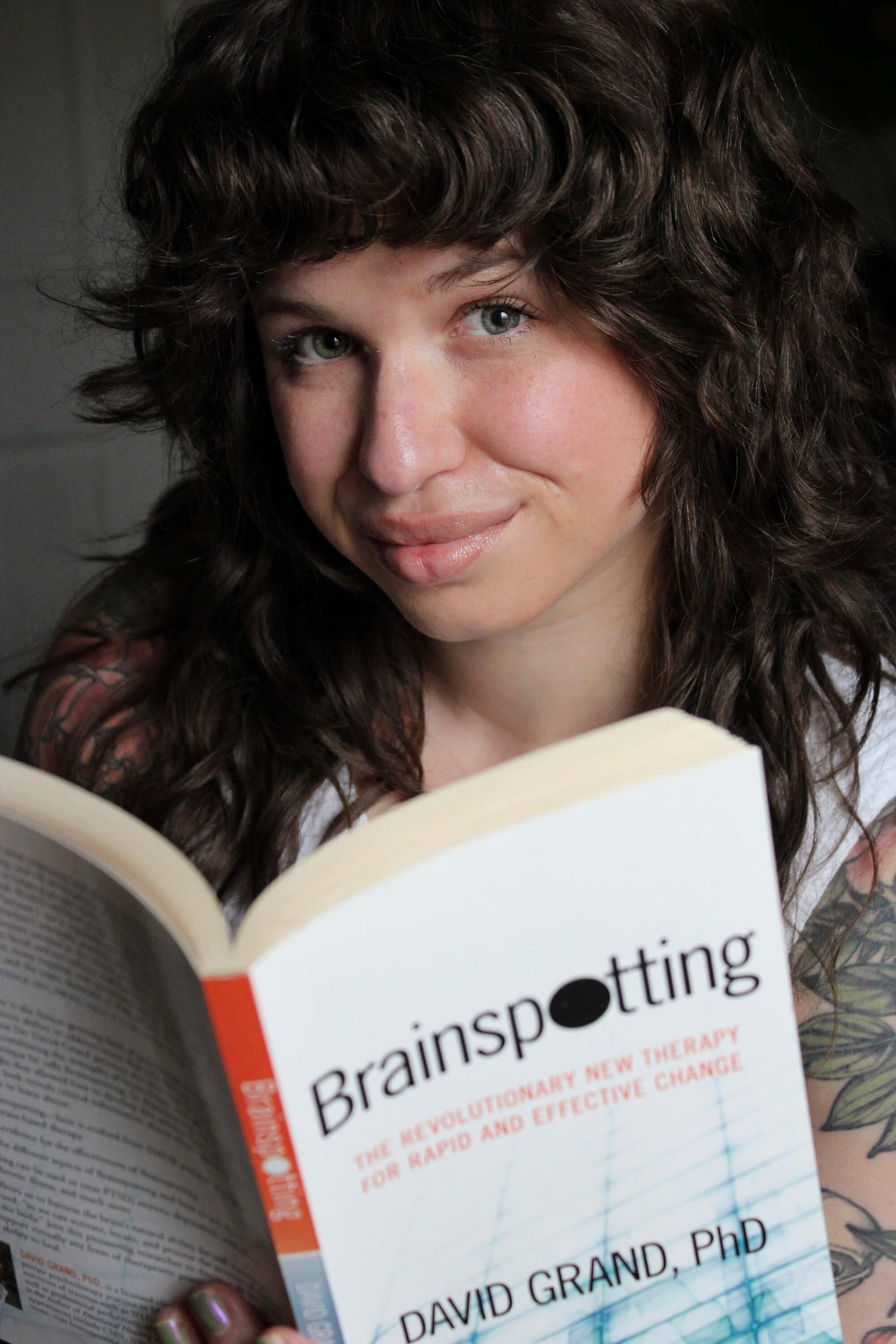 A woman with curly brown hair and tattoos holding a book titled 'Brainspotting' by David Grand, PhD, smiling at the camera.