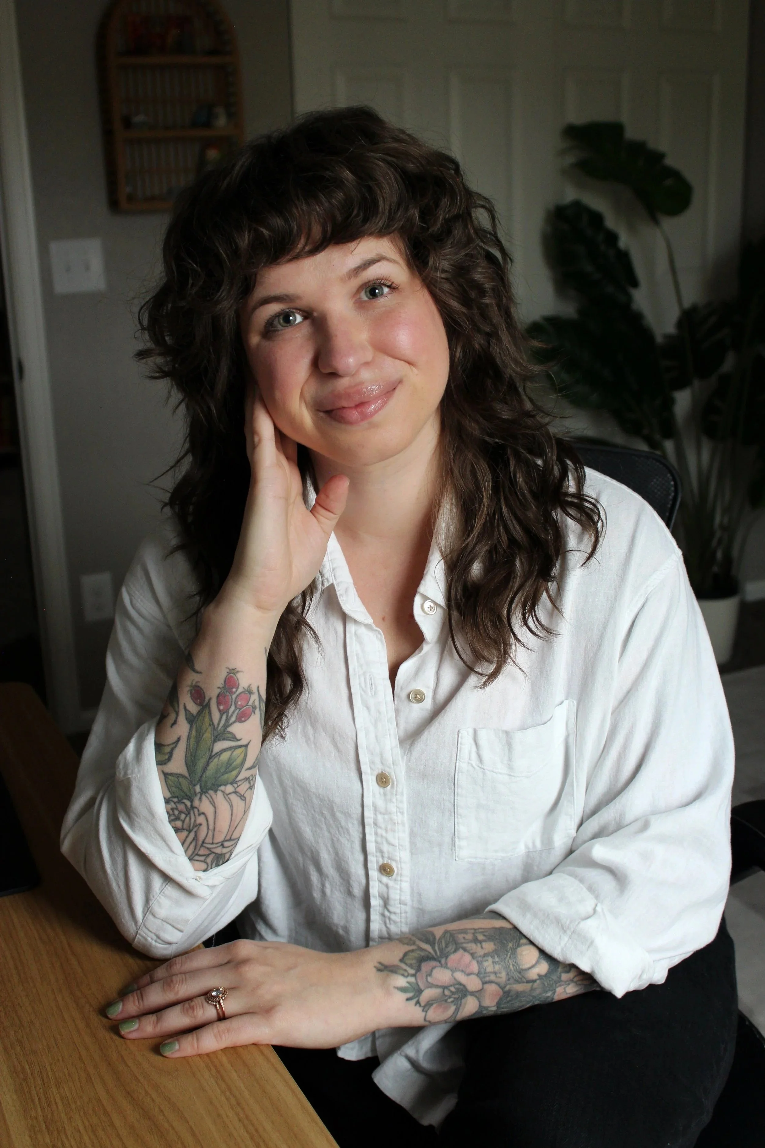 A woman with curly brown hair and tattoos on her arms, wearing a white button-up shirt, sitting at a wooden table with her hand resting on her cheek, smiling softly.