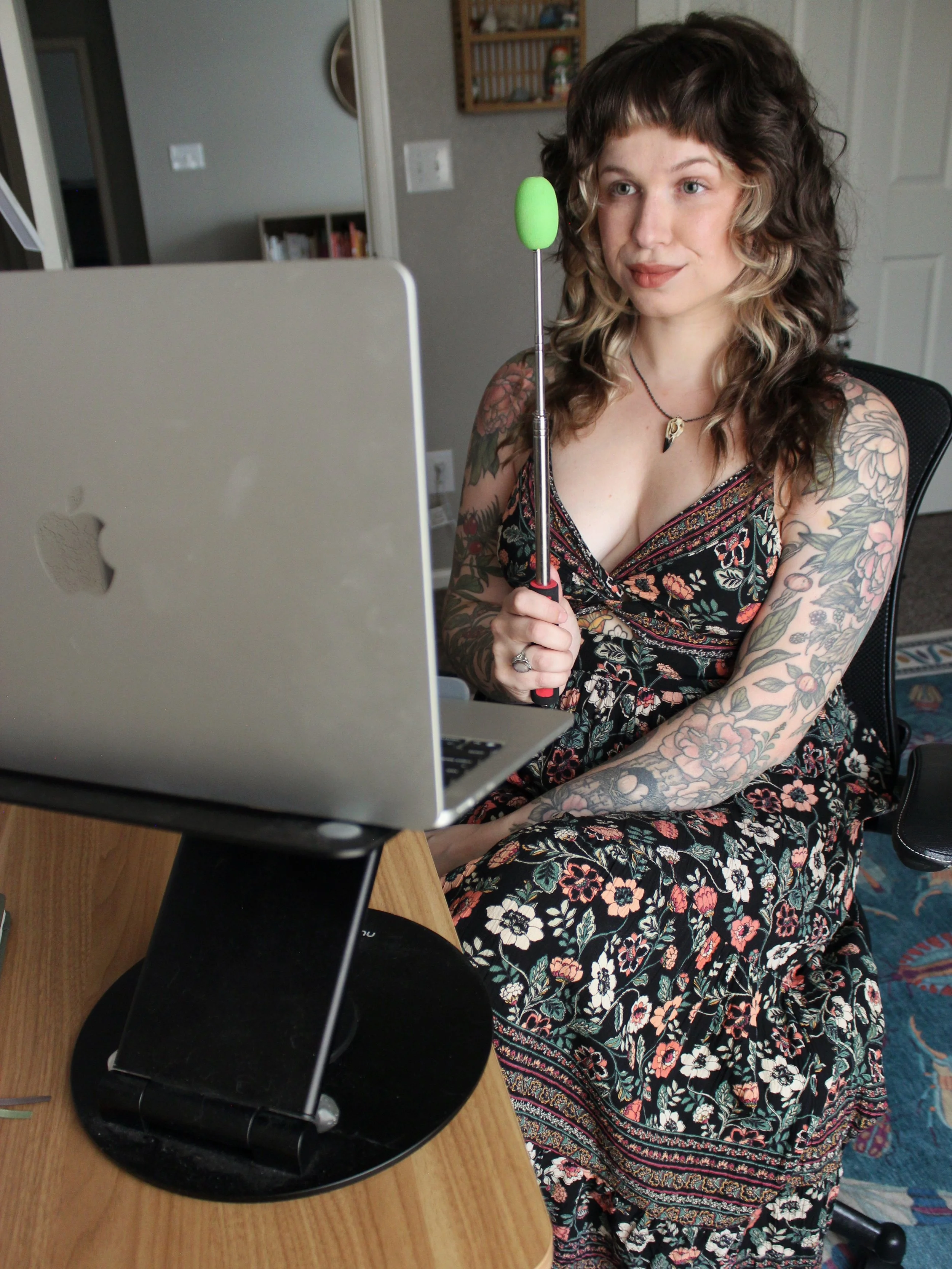 A woman with tattoos on her arms and chest, wearing a floral dress, is sitting in front of a laptop, holding a green club-shaped object, in a home setting with books and a mirror in the background.