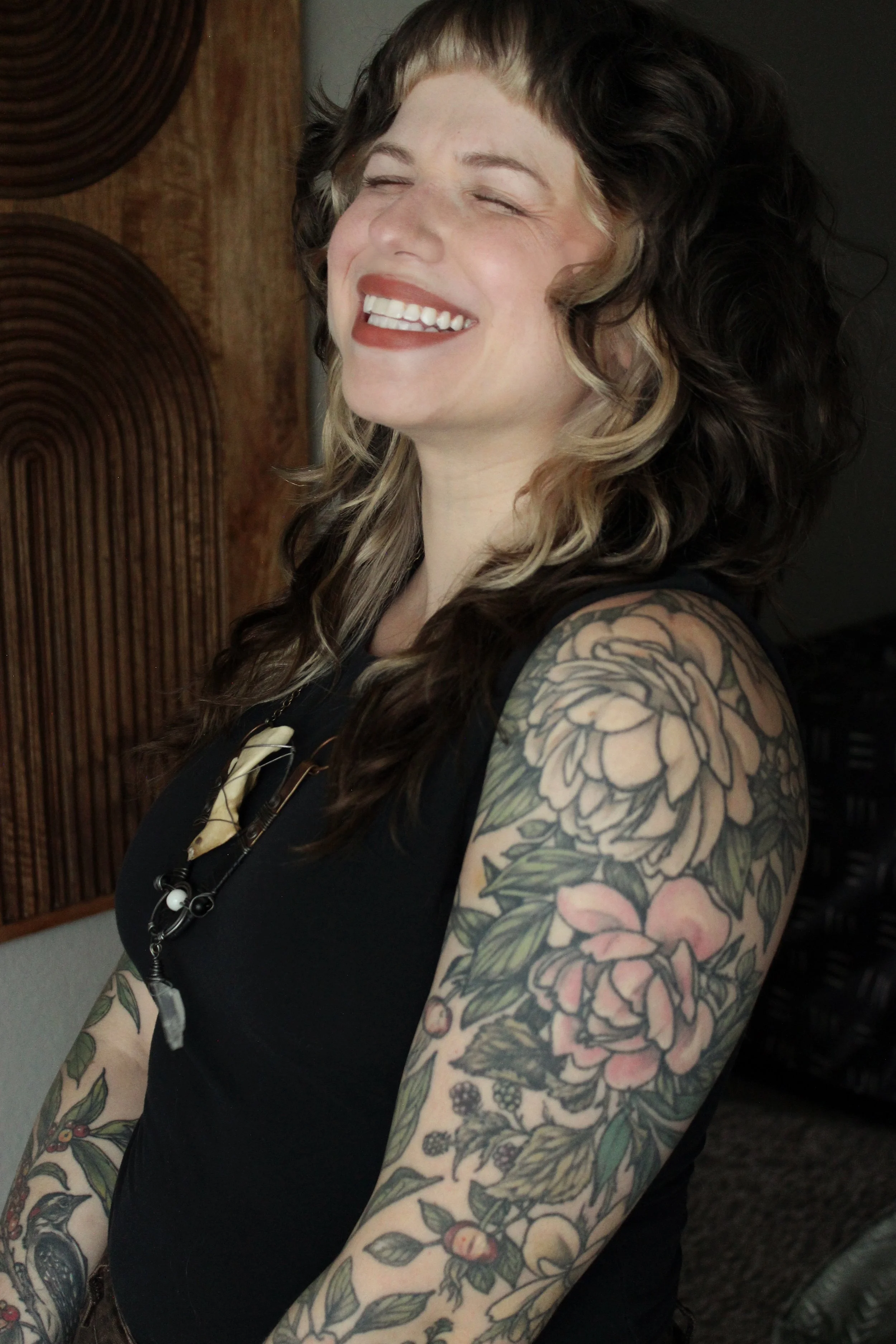 A woman with curly black hair and blonde highlights, smiling with eyes closed, showing facial tattoos on arms and shoulders, wearing a black sleeveless top, standing indoors with a wooden wall and dark background.
