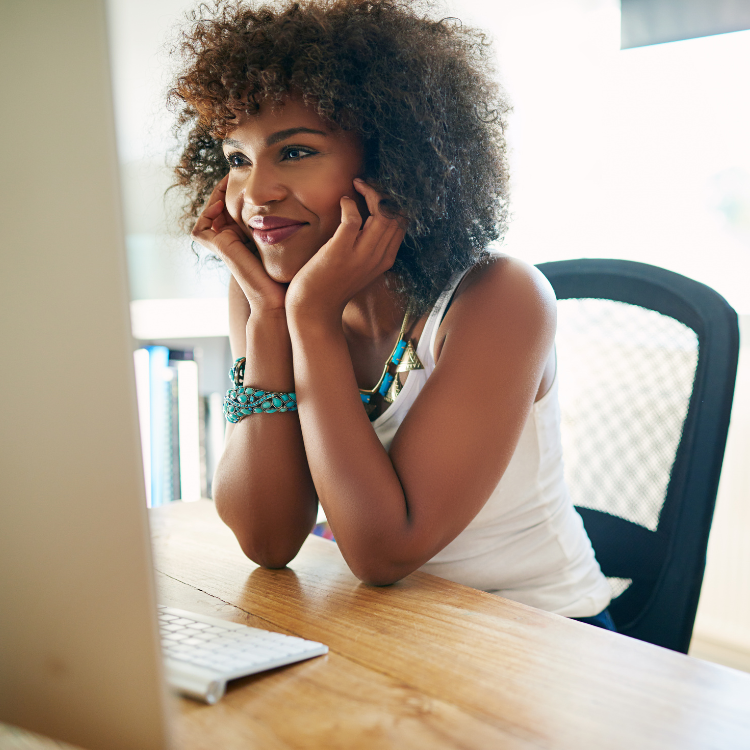 A woman with curly hair smiling while looking at her computer screen, sitting at a wooden desk with a keyboard in front of her.