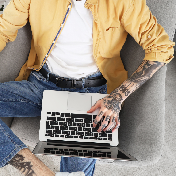 Person sitting on a gray sofa using a laptop, wearing a yellow jacket, white shirt, and blue jeans, with tattoos on their arm and hand.