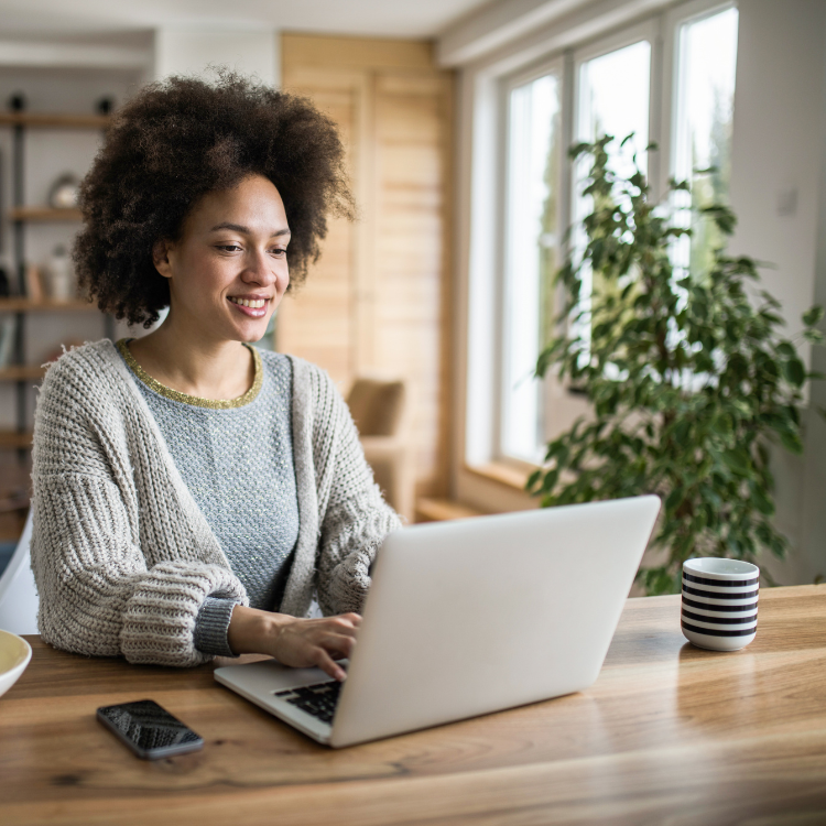 A woman with curly hair smiling while working on a laptop at a wooden table in a bright room with large windows and indoor plants.