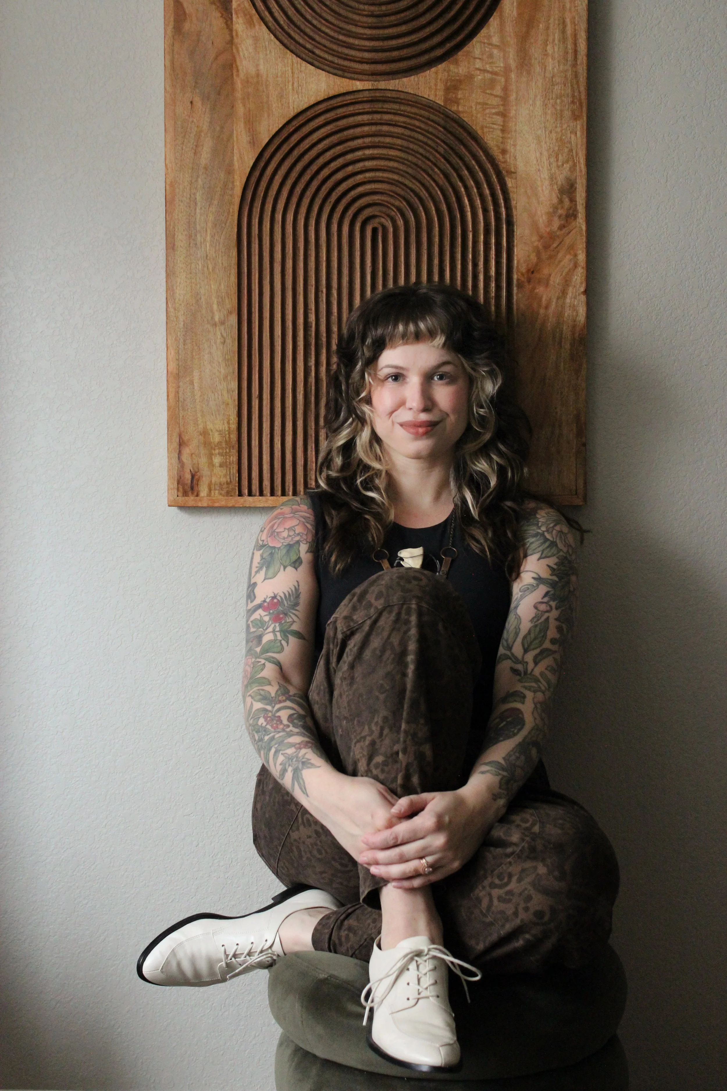 A woman with tattoos on her arms and wavy dark hair with blonde highlights, sitting cross-legged on a black ottoman against a plain wall with a wooden decorative piece behind her. She is wearing a black top, brown patterned pants, and white shoes, and is smiling at the camera.